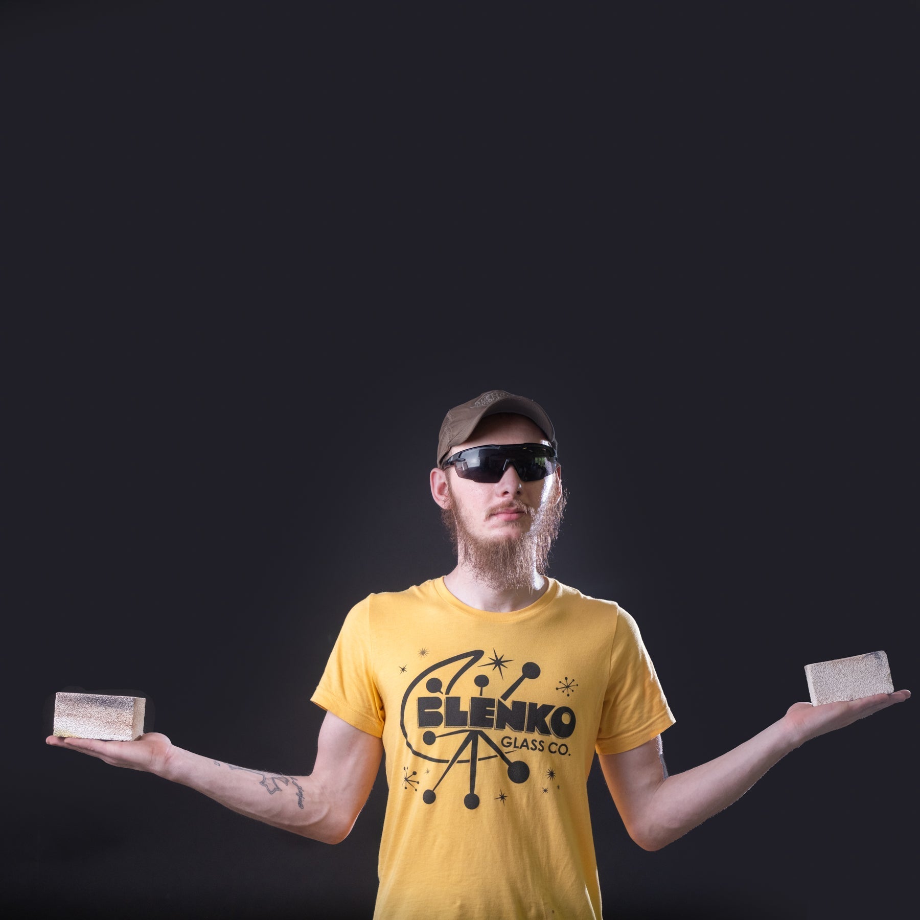 Man wearing a yellow t-shirt with 'Blenko Glass Co.' logo, holding two glass blocks against a black background.