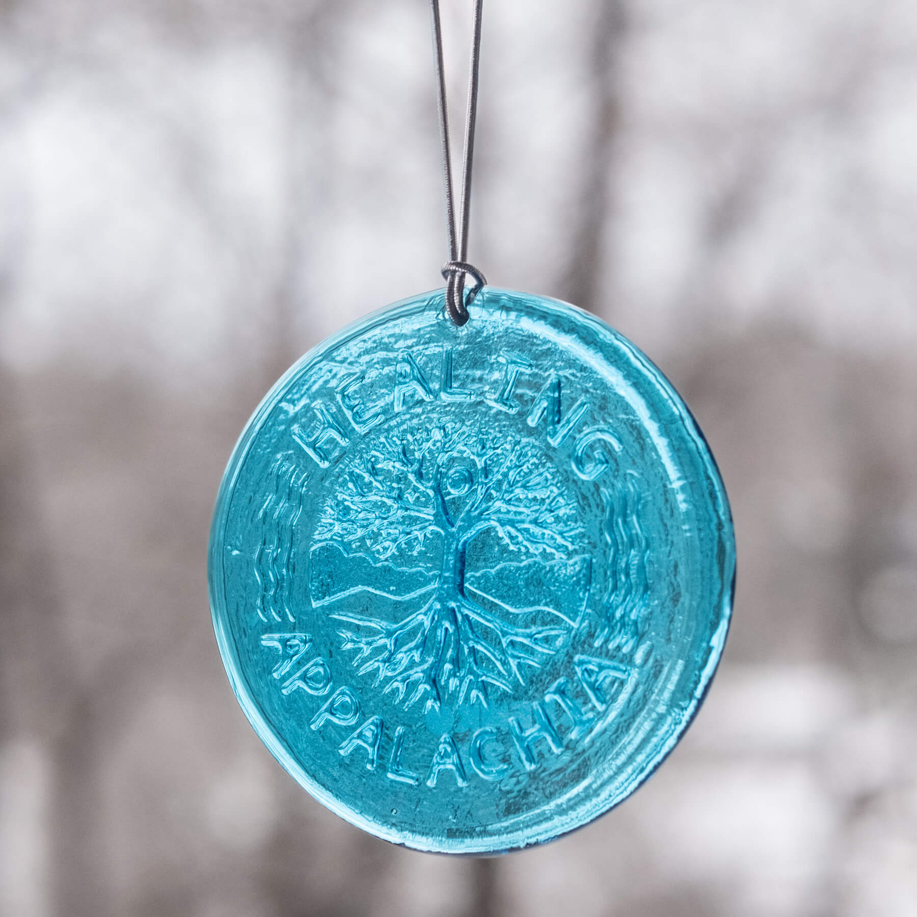 A round glass suncatcher with a turquoise tint, featuring an embossed design of a dragon and the text 'Healing Appalachia', hanging by a string.