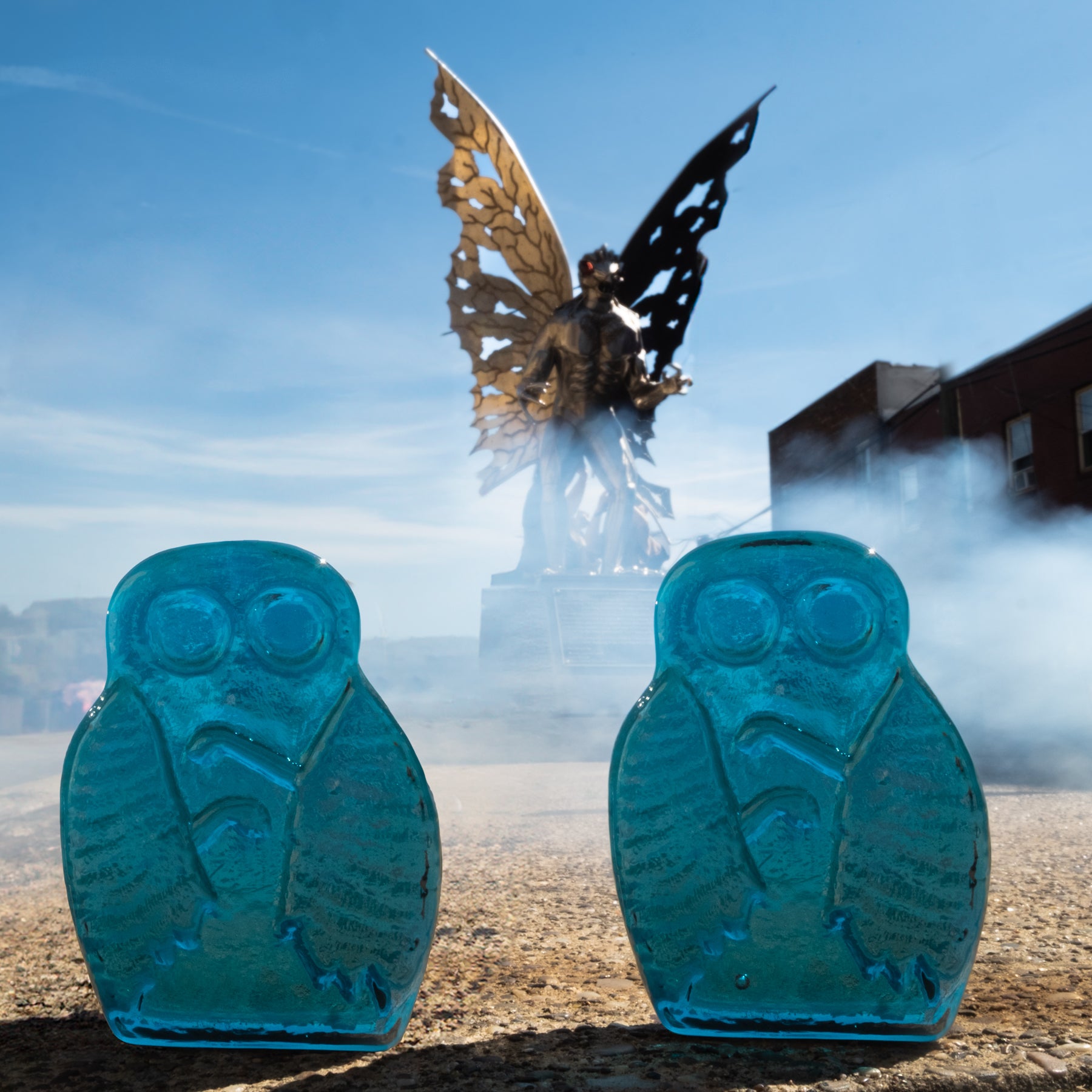 Two blue owl-shaped objects in front of a large butterfly sculpture with a cityscape in the background.