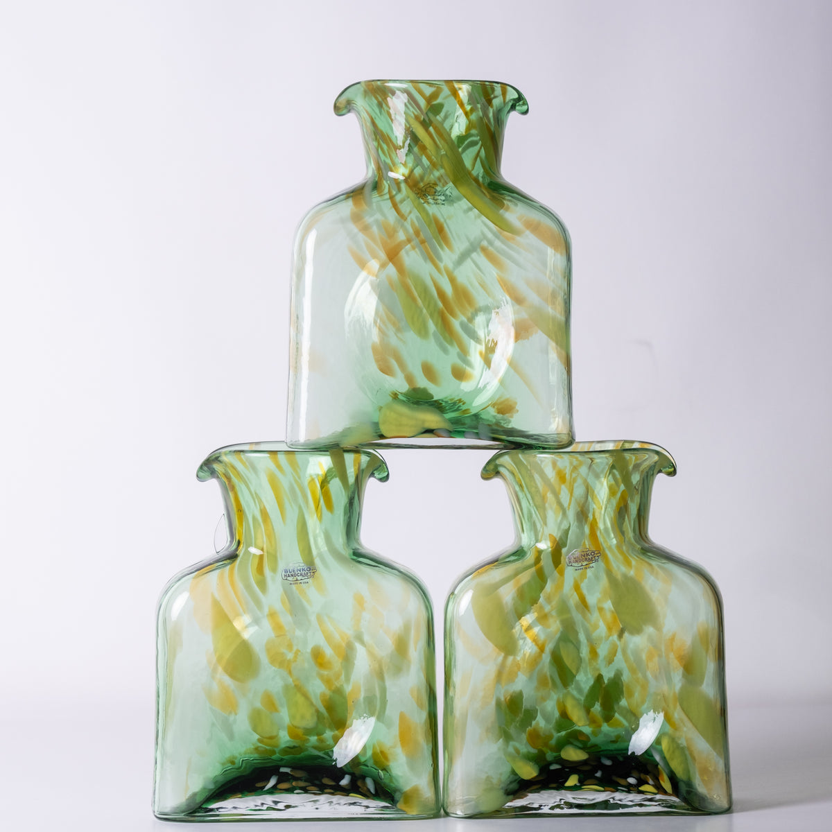 Three tall, narrow glass bottles with flared rims, stacked in a pyramid arrangement. The bottles are made of transparent light green glass with abstract yellow and olive-green marbled swirls, set against a plain light gray background.