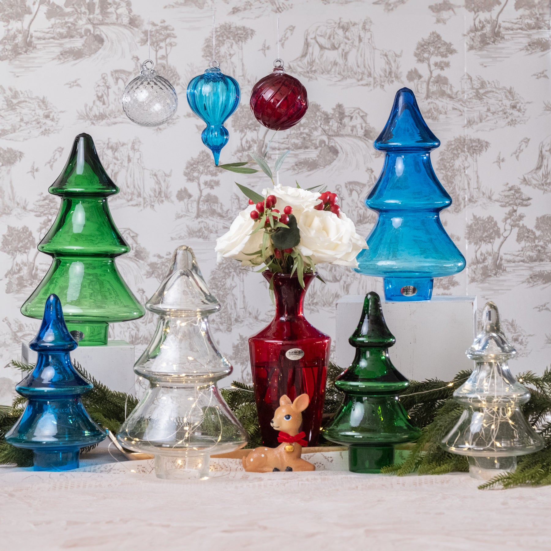 Decorative glass Christmas trees and ornaments on a table with a floral arrangement against a patterned wall.