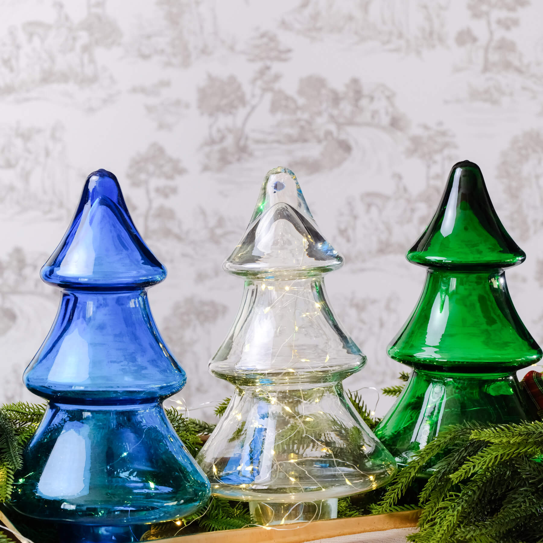 Three glass Christmas trees in blue, clear, and green on a bed of greenery with a textured wall background.