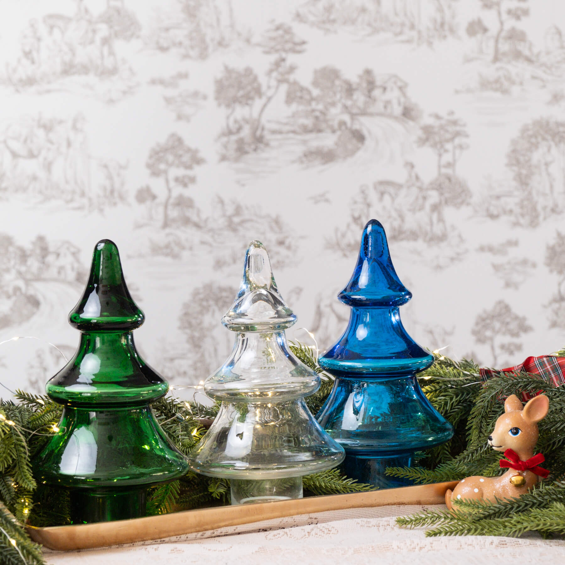 Three glass Christmas trees in green, clear, and blue on a wooden tray with a white textured background.