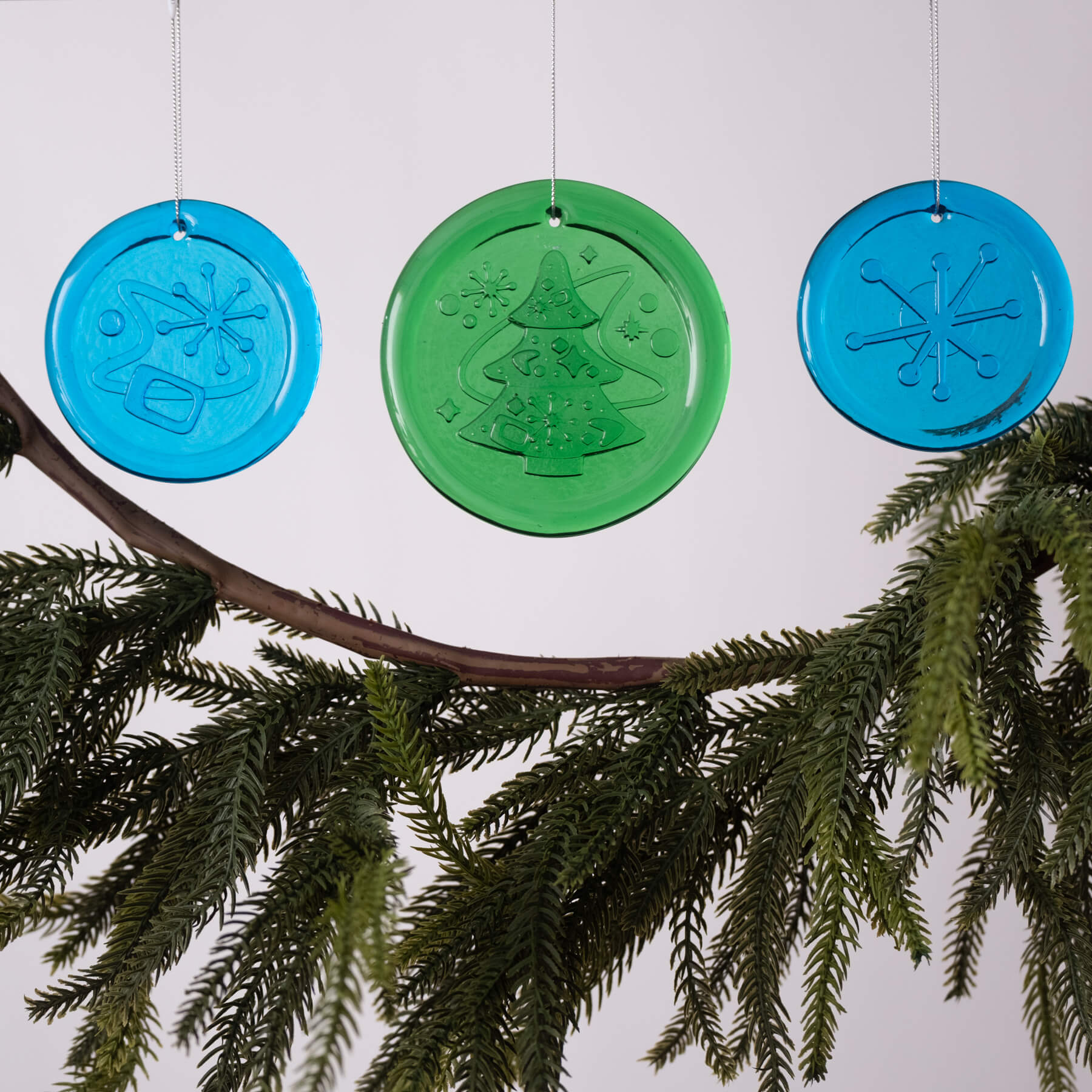 Three circular Christmas ornaments, two blue and one green, hanging on a branch against a white background.