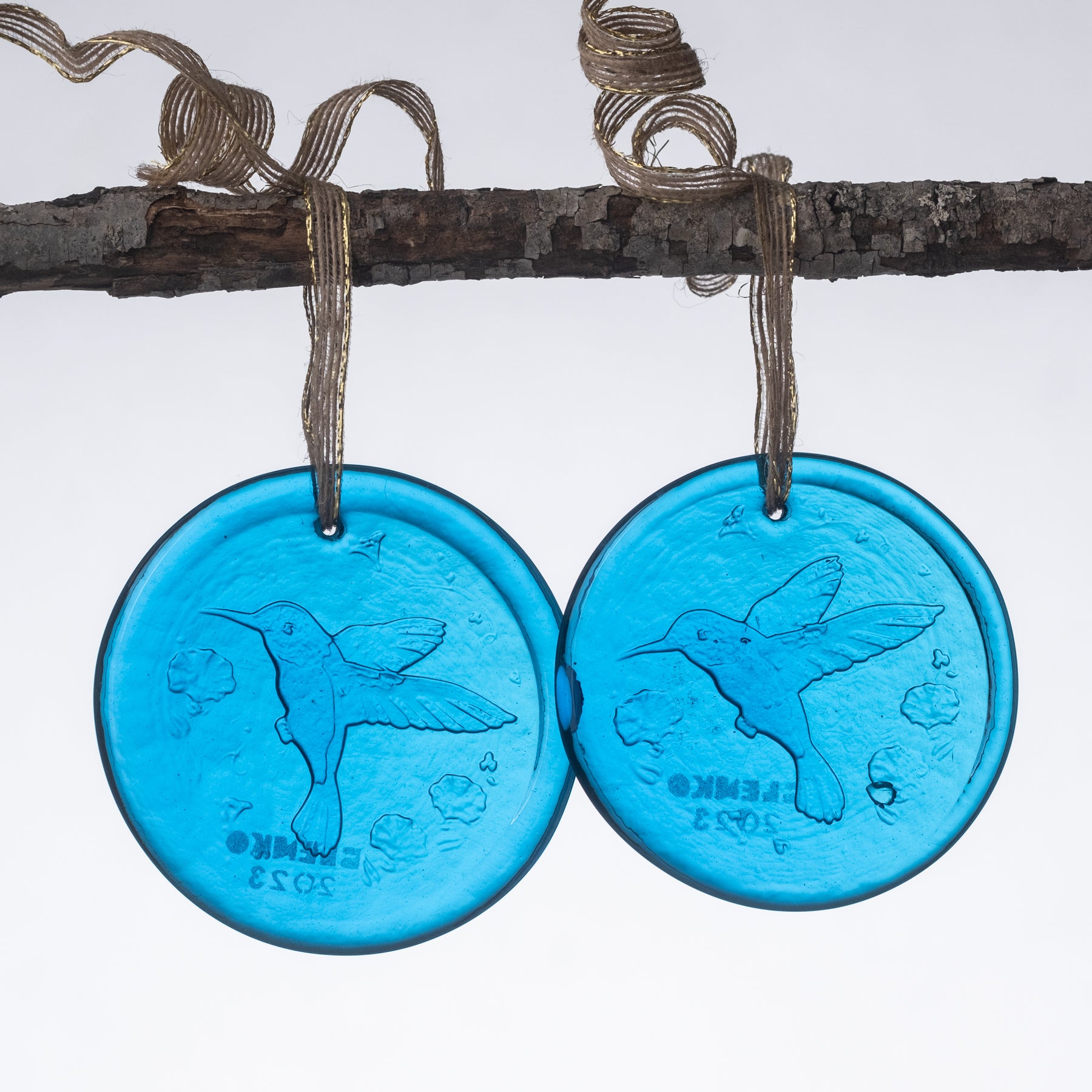 Two round glass suncatchers with a turquoise blue hue, featuring an engraved design of a hummingbird, flowers, and possibly a sun. The suncatcher has a metallic loop at the top for hanging.
