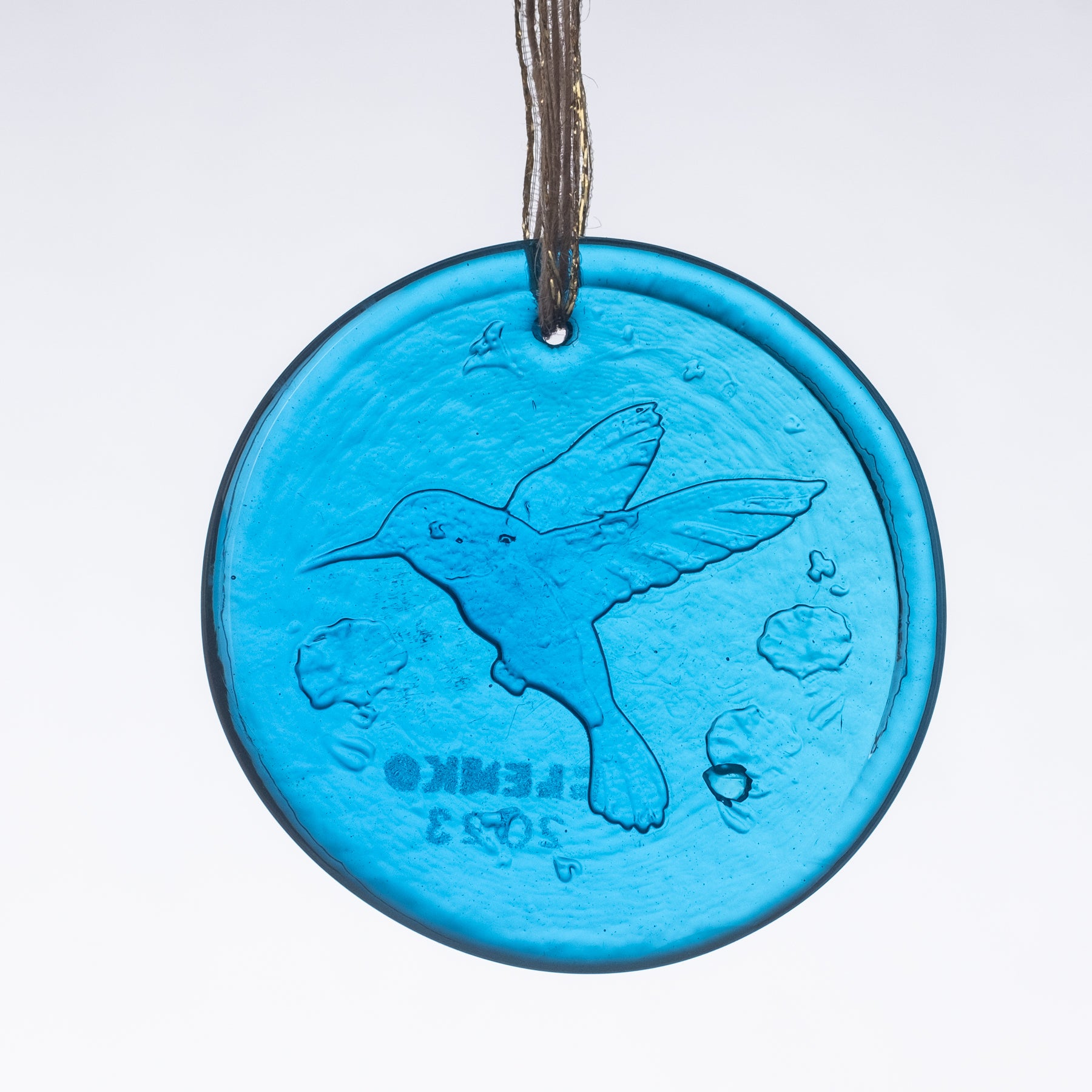 A round glass suncatcher with a turquoise blue hue, featuring an engraved design of a hummingbird, flowers, and possibly a sun. The suncatcher has a metallic loop at the top for hanging.