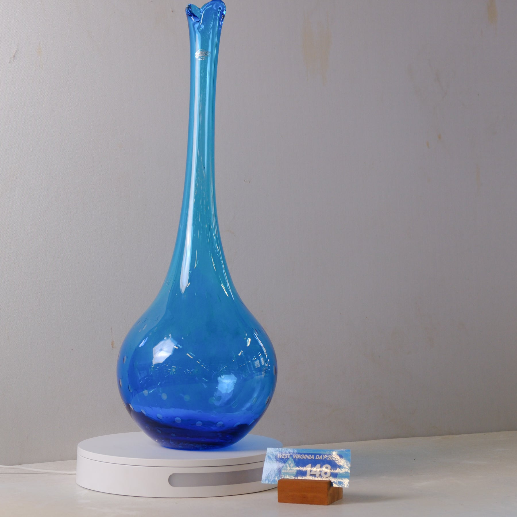 A cobalt blue glass vase with a flared neck and bulbous base, displayed on a white circular light box with a small price tag in front, against a plain light gray wall.
