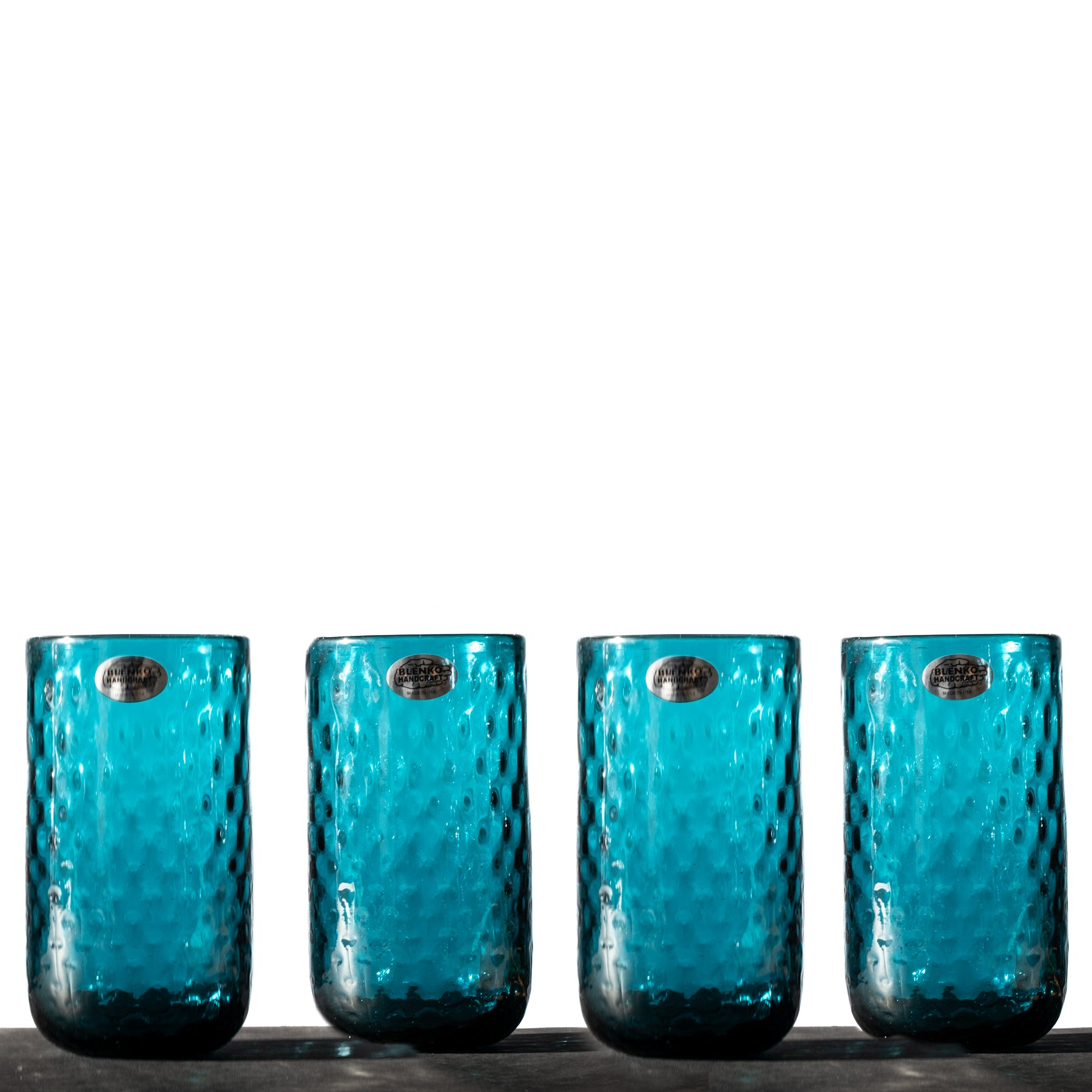 A set of four hand-blown blue glass tumblers with a diamond optic pattern, displayed side by side.