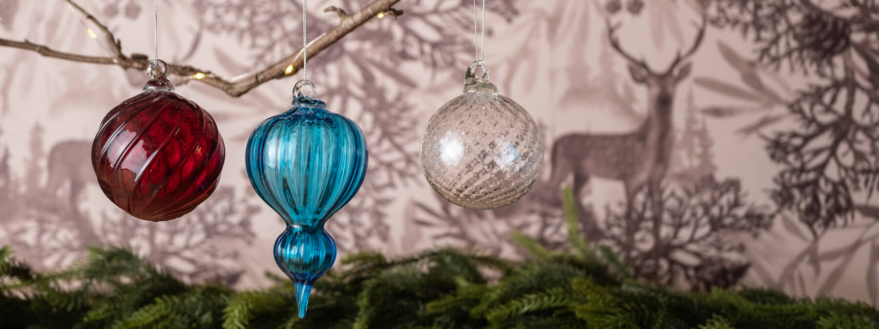 Three glass Christmas ornaments hanging on a branch with a deer and floral patterned background.