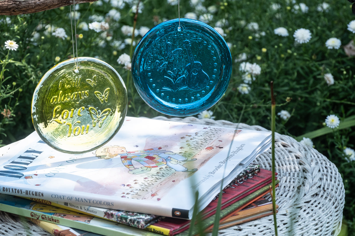 Two decorative glass wind chimes, one translucent yellow-green and one opaque teal blue, hanging outdoors on a stack of books in a garden setting.