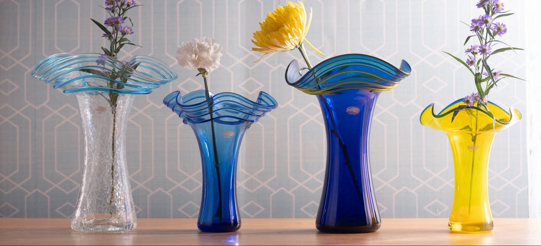 Set of four colorful glass vases with flowers against a patterned wall.