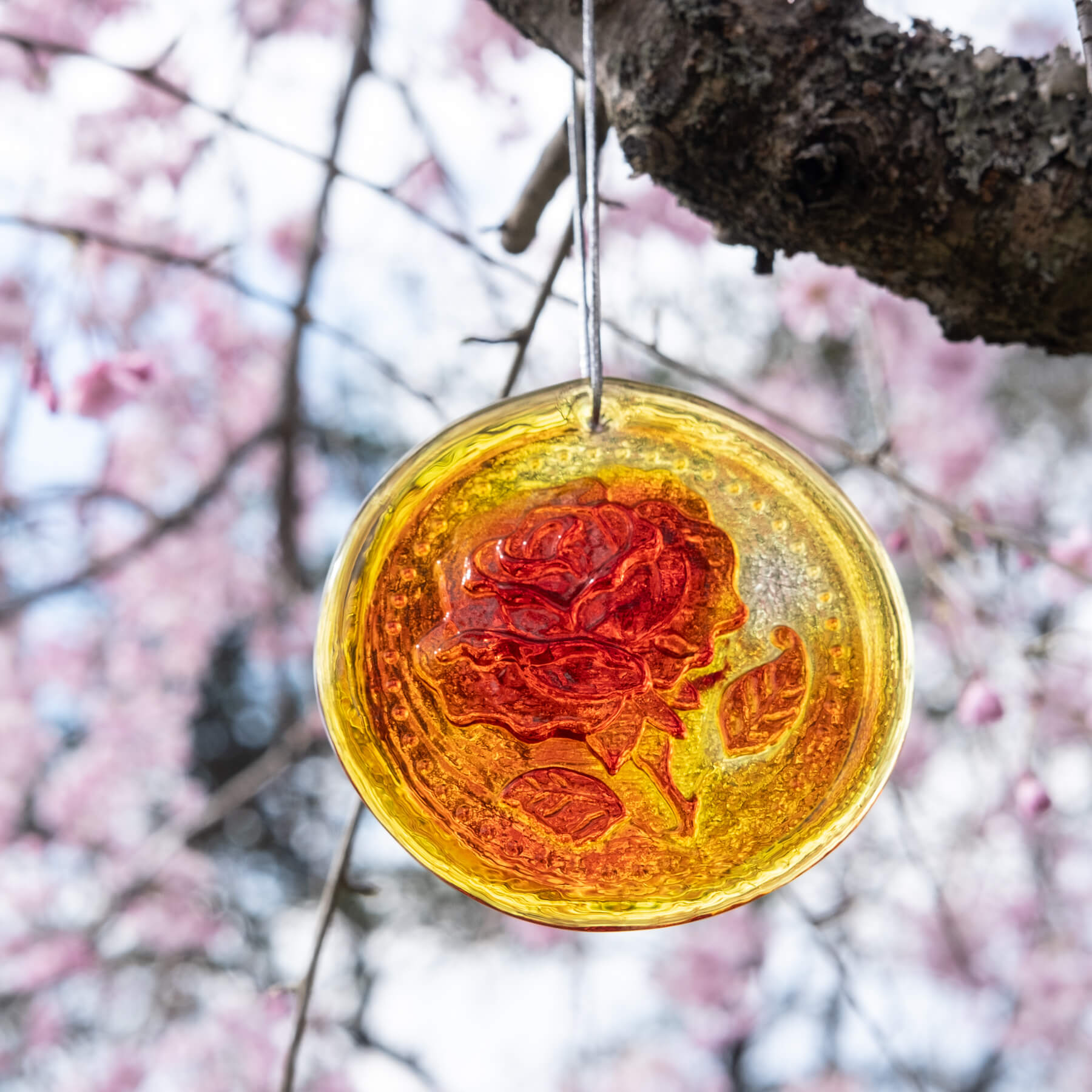 A handcrafted glass suncatcher with a rose design, in orange and yellow hues, hanging by a string against a blurred background of pink flowers and greenery.