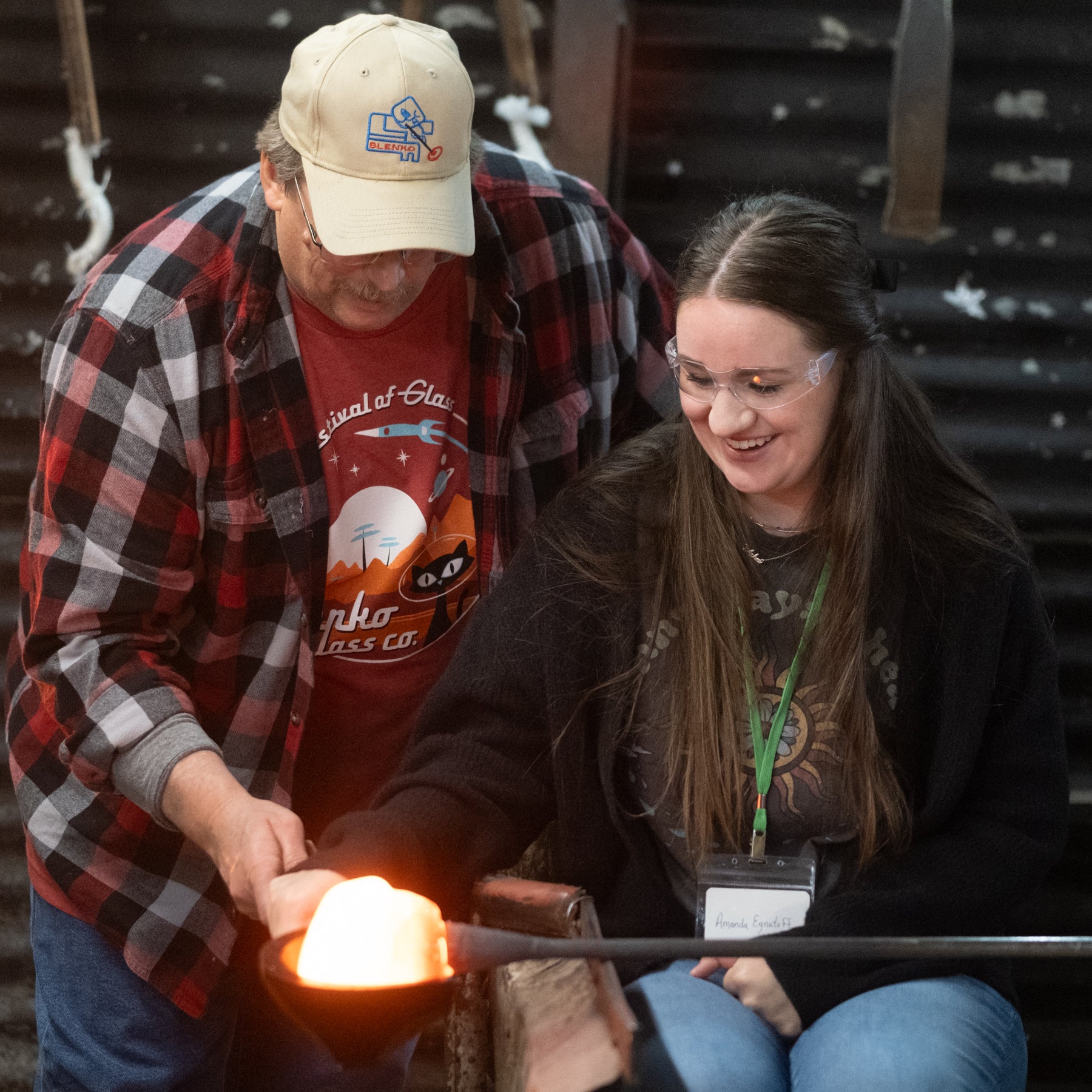 A person participating in a glassblowing experience, shaping a hot piece of glass with the assistance of an artisan.