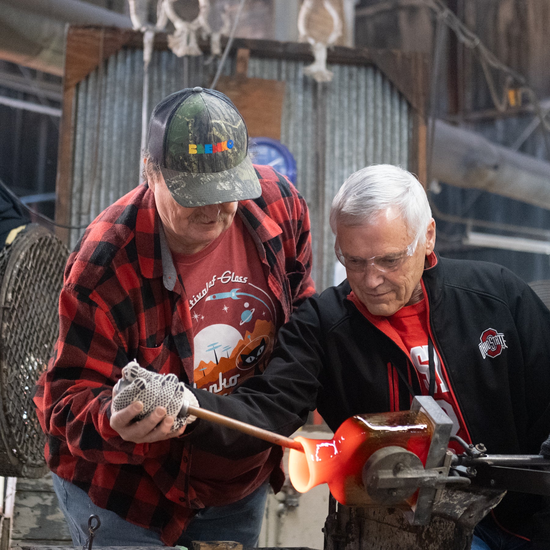 Two men working with a hot object on an anvil, one wearing a cap and plaid shirt, the other in a black jacket.