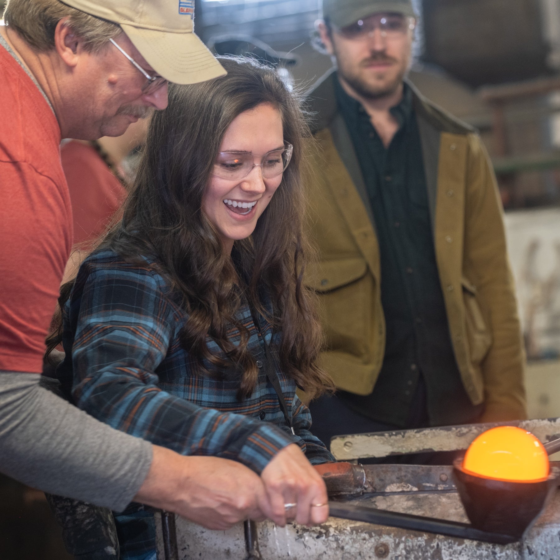 Woman working with hot metal in a workshop setting, with two men observing.