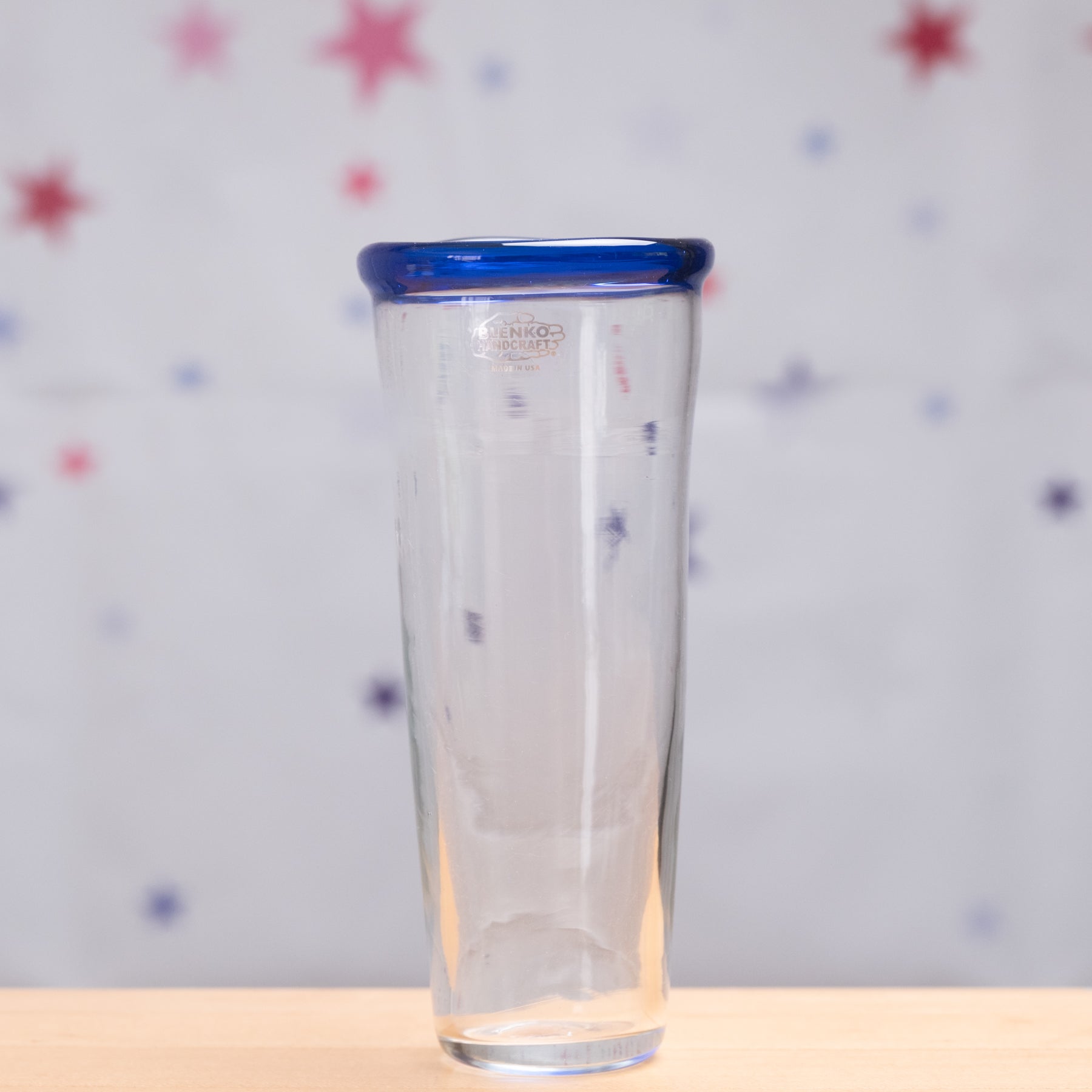 Clear glass tumbler with a blue rim on a light wooden surface and star-patterned background
