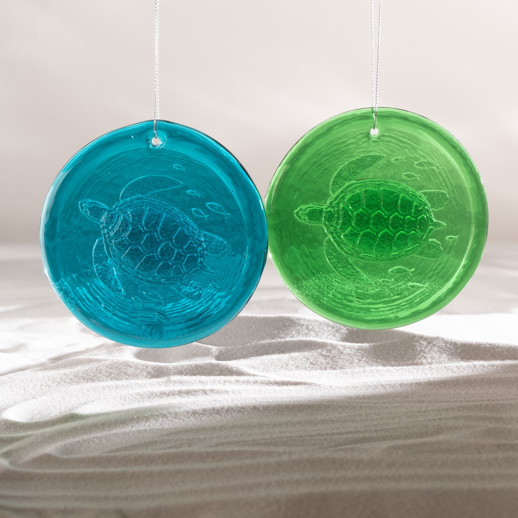 Two round hanging glass ornaments with embossed sea turtle designs, one translucent blue and one translucent green, displayed against a soft light background.