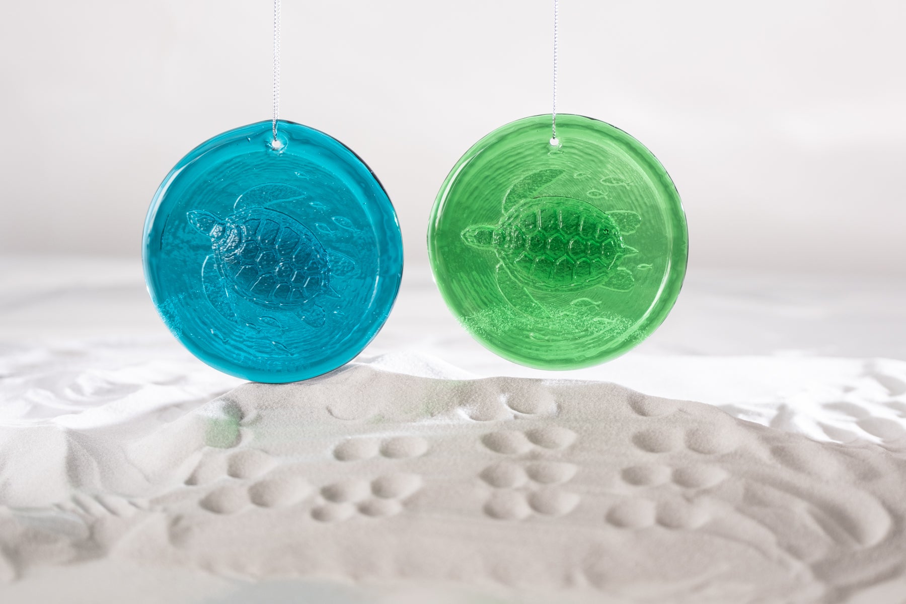 Two round translucent turtle-shaped ornaments, one blue and one green, hanging against a white background above a white textured surface.