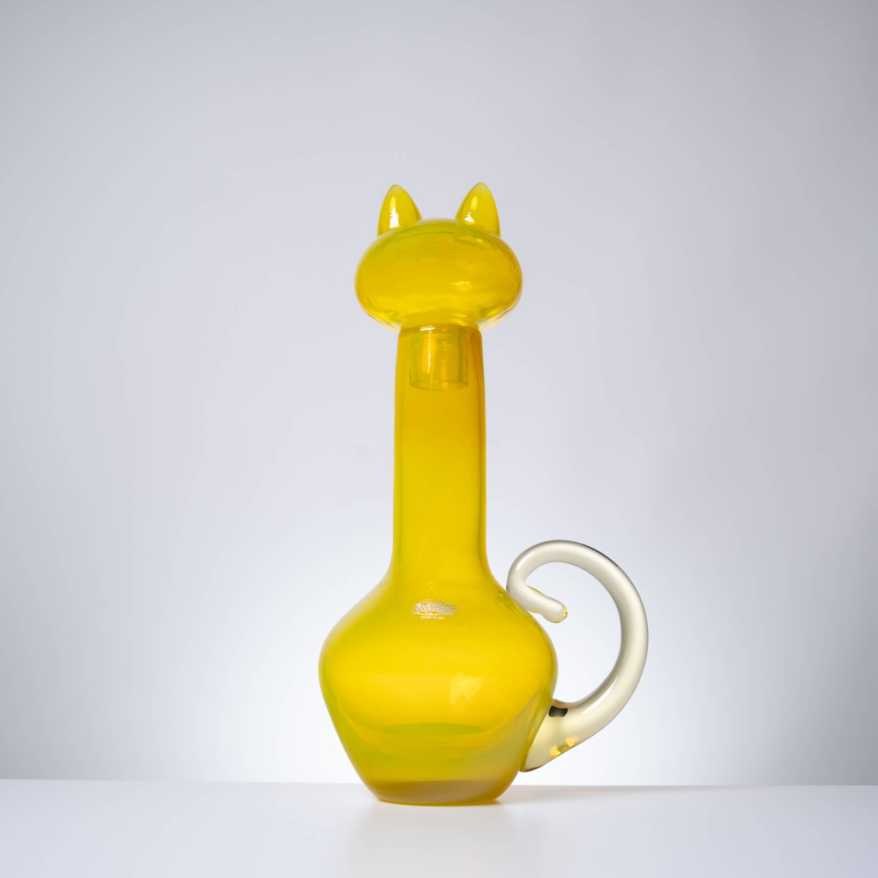A translucent yellow glass bottle shaped like a cat, featuring a long neck, pointed ears, a curved handle, and a small white accent near the base, photographed against a light gray studio background.