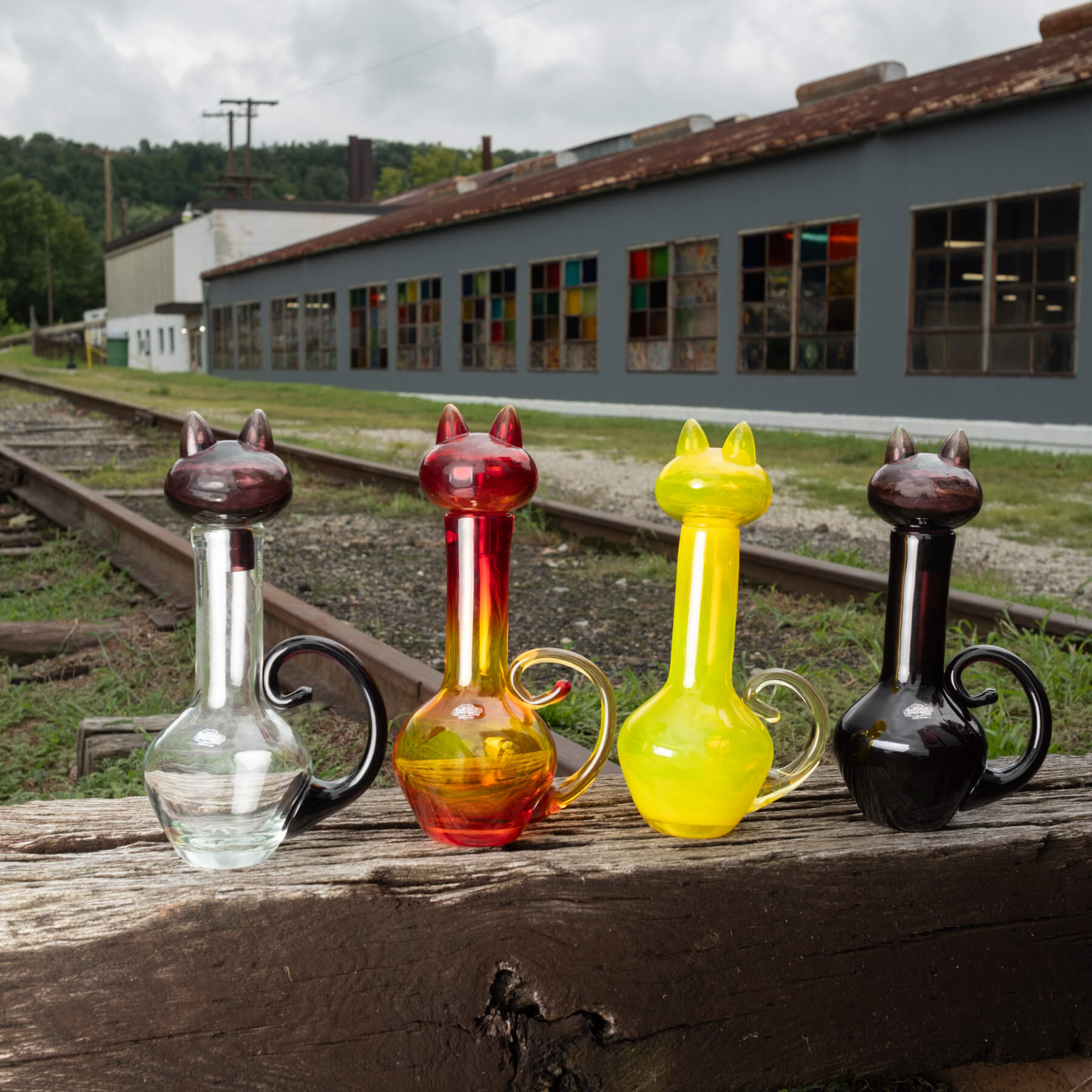 Four cat-shaped glass decanters with cat heads and handles are lined up on a weathered wooden beam outdoors near railroad tracks and a gray warehouse building.