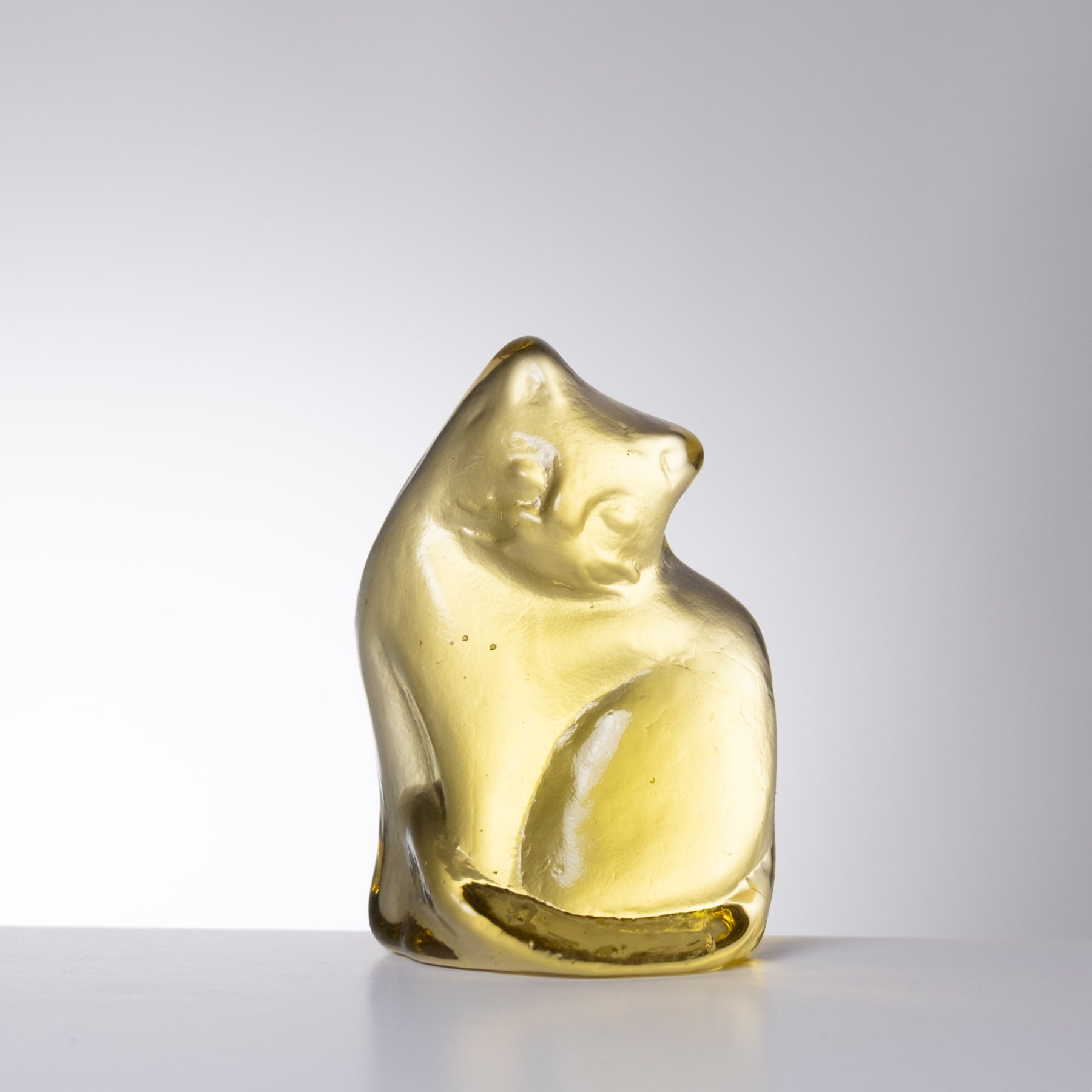 A small translucent amber-yellow glass figurine of a curled-up cat, standing on a white surface against a light gray gradient background.