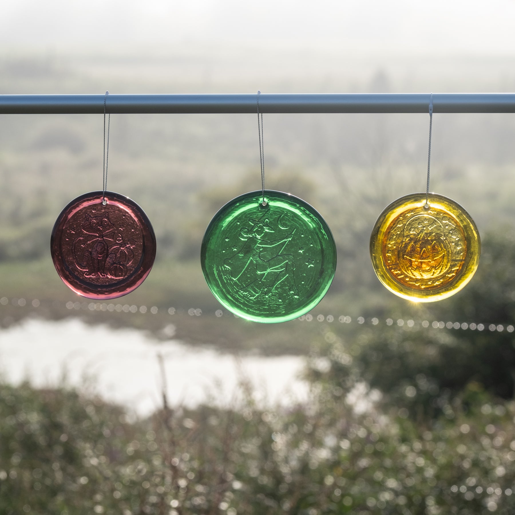 Three circular hanging glass ornaments in red, green, and yellow are suspended from a metal rod outdoors, with a blurred landscape in the background.