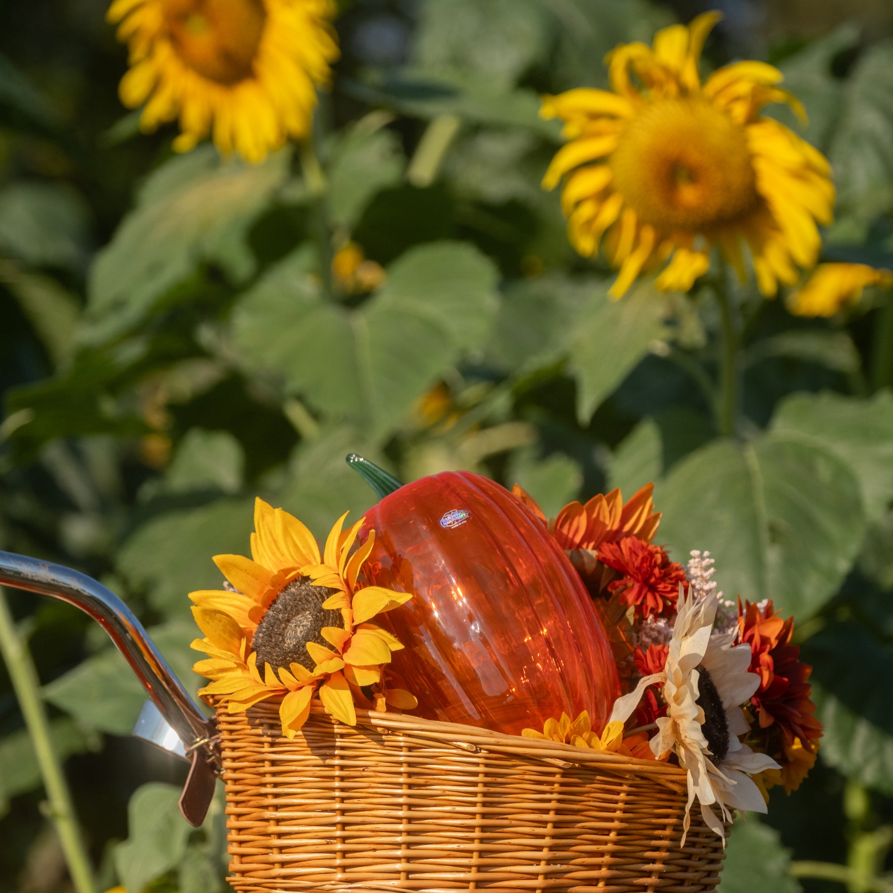 A wicker basket filled with a glossy orange-red decorative pumpkin and sunflowers, set against a blurred background of real sunflowers and green foliage.