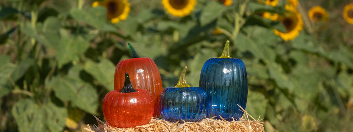 Four ribbed glass pumpkins in red, orange, blue, and dark blue, arranged on a bed of straw with a blurred background of sunflowers.