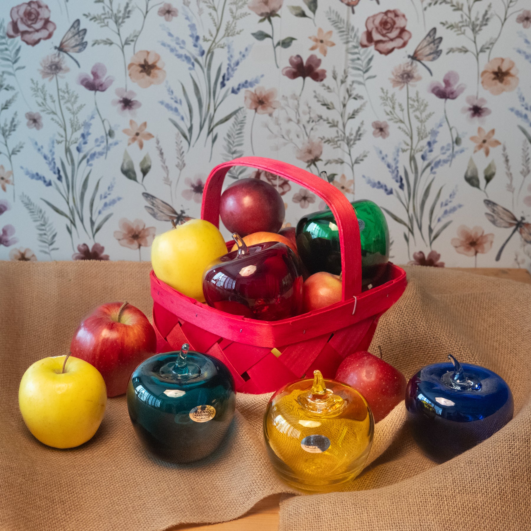 Colorful glass apples with a red basket filled with apples against a floral wallpaper background.