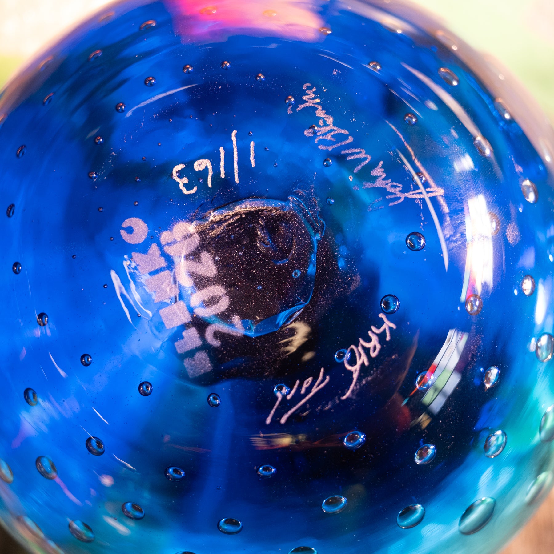 A close-up of a blue glass ornament or decorative glass sphere with a bubbled texture, featuring white handwritten signatures and dates on the inside.