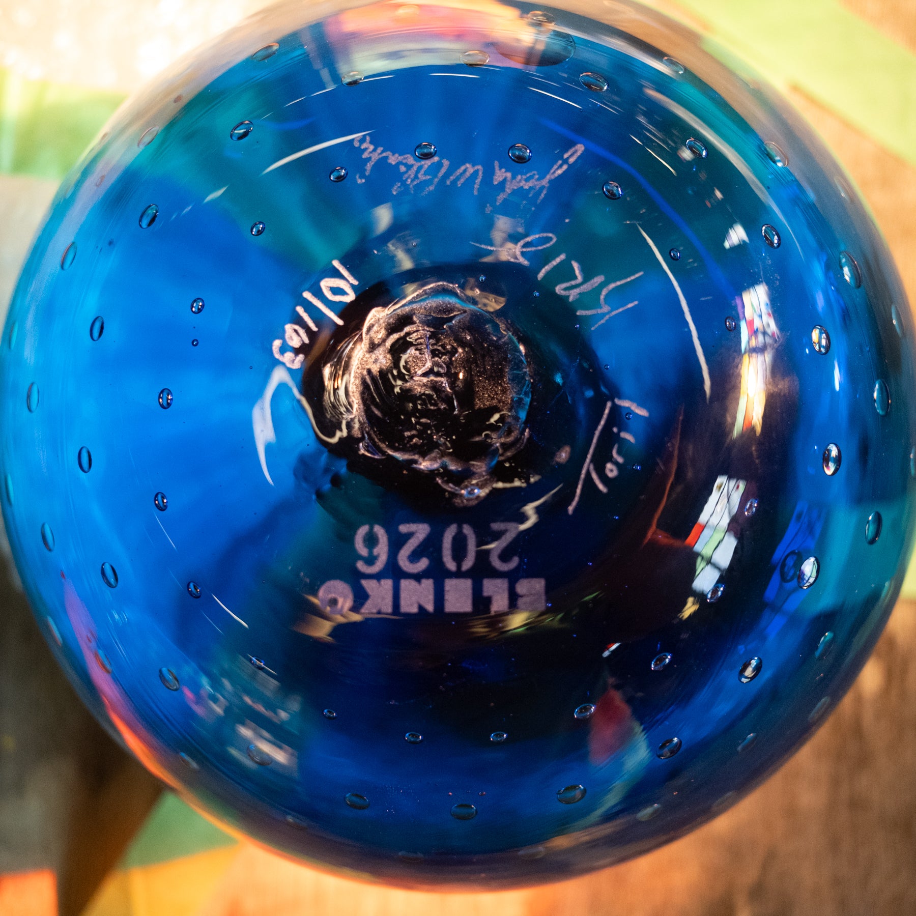 A close-up of a blue glass bottle or container viewed from the bottom, showing a dark stopper, small bubble-like perforations around the rim, and white printed text including '2026' and 'BLENK'.