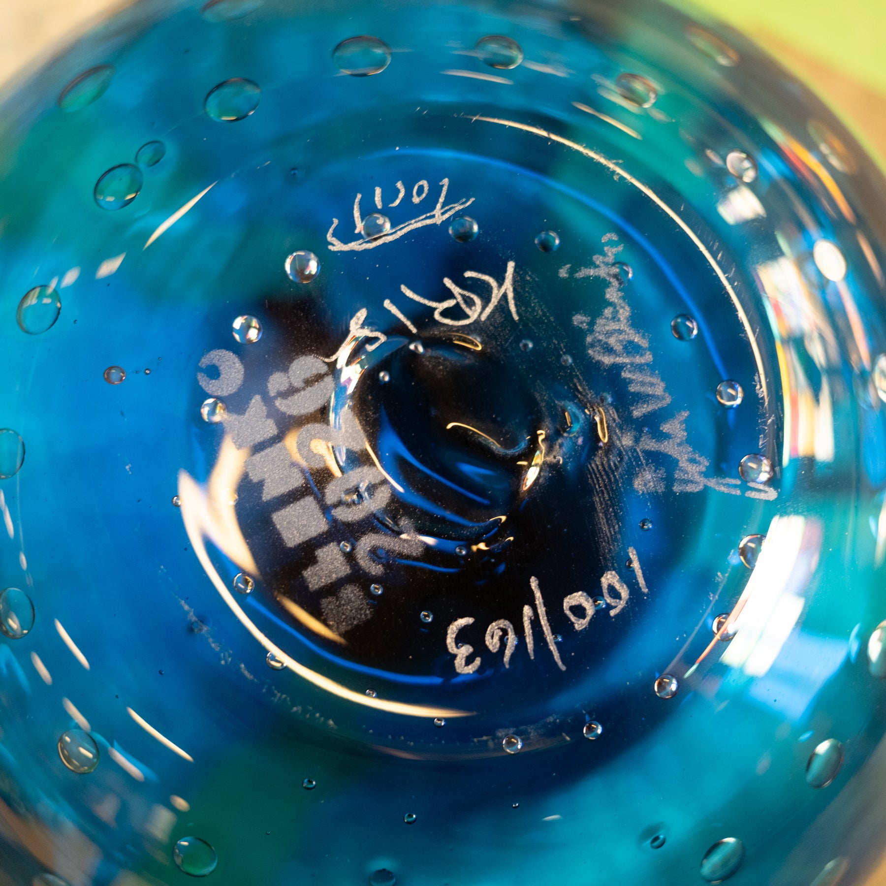 Close-up of the bottom of a blue glass bowl or vase, showing etched maker's marks, a date stamp reading 1963, and small air bubbles embedded in the glass.