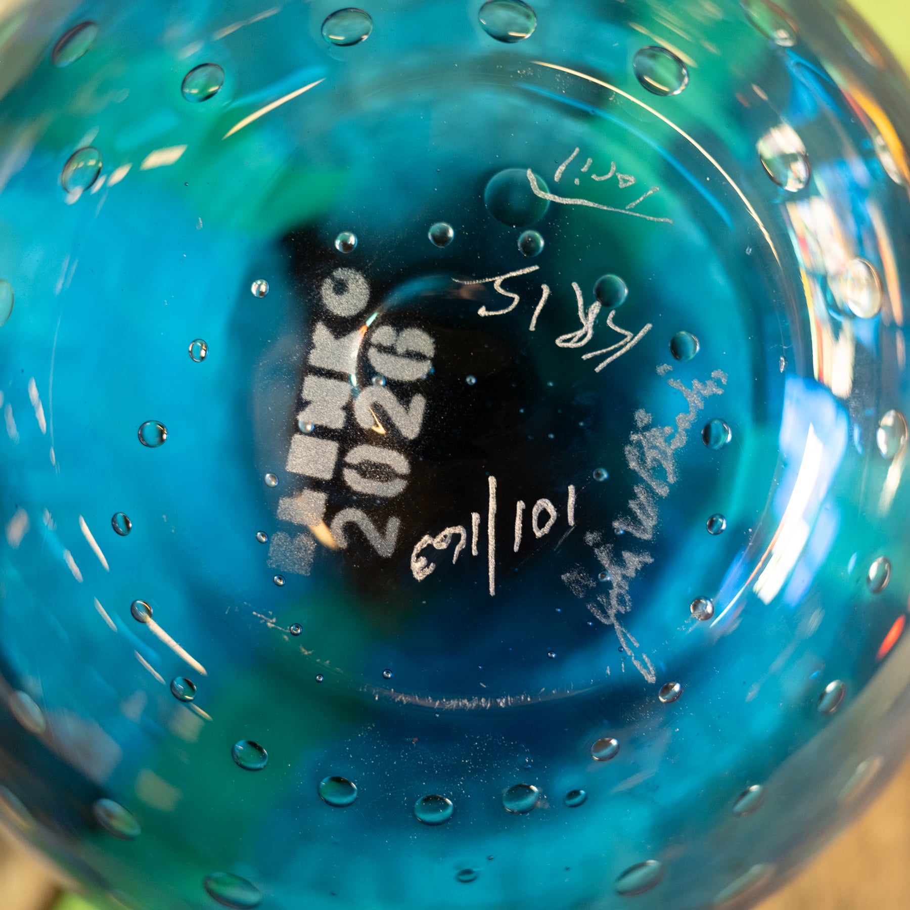 Close-up of the bottom of a hand-blown glass vessel, showing a vibrant blue and green gradient with white bubble details and white printed markings including 'SILKNO 2026' and handwritten numbers.