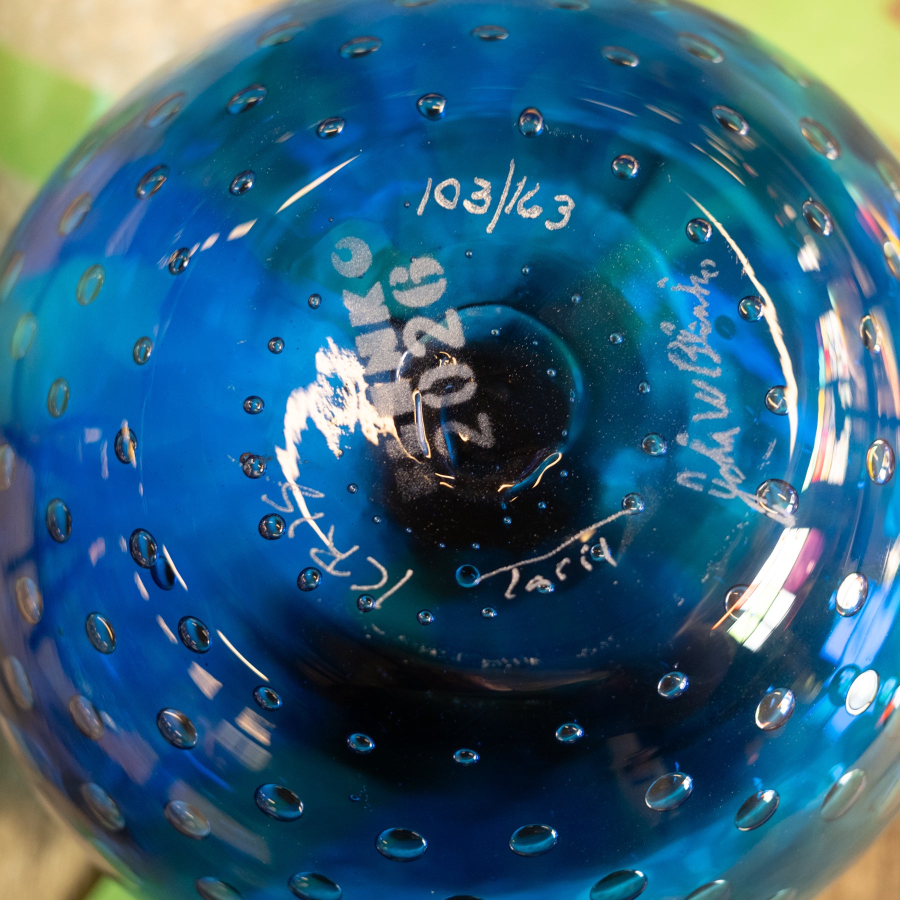 Close-up of a blue glass bowl or vase with a perforated bubble pattern, showing white handwritten markings and signatures on the bottom surface.