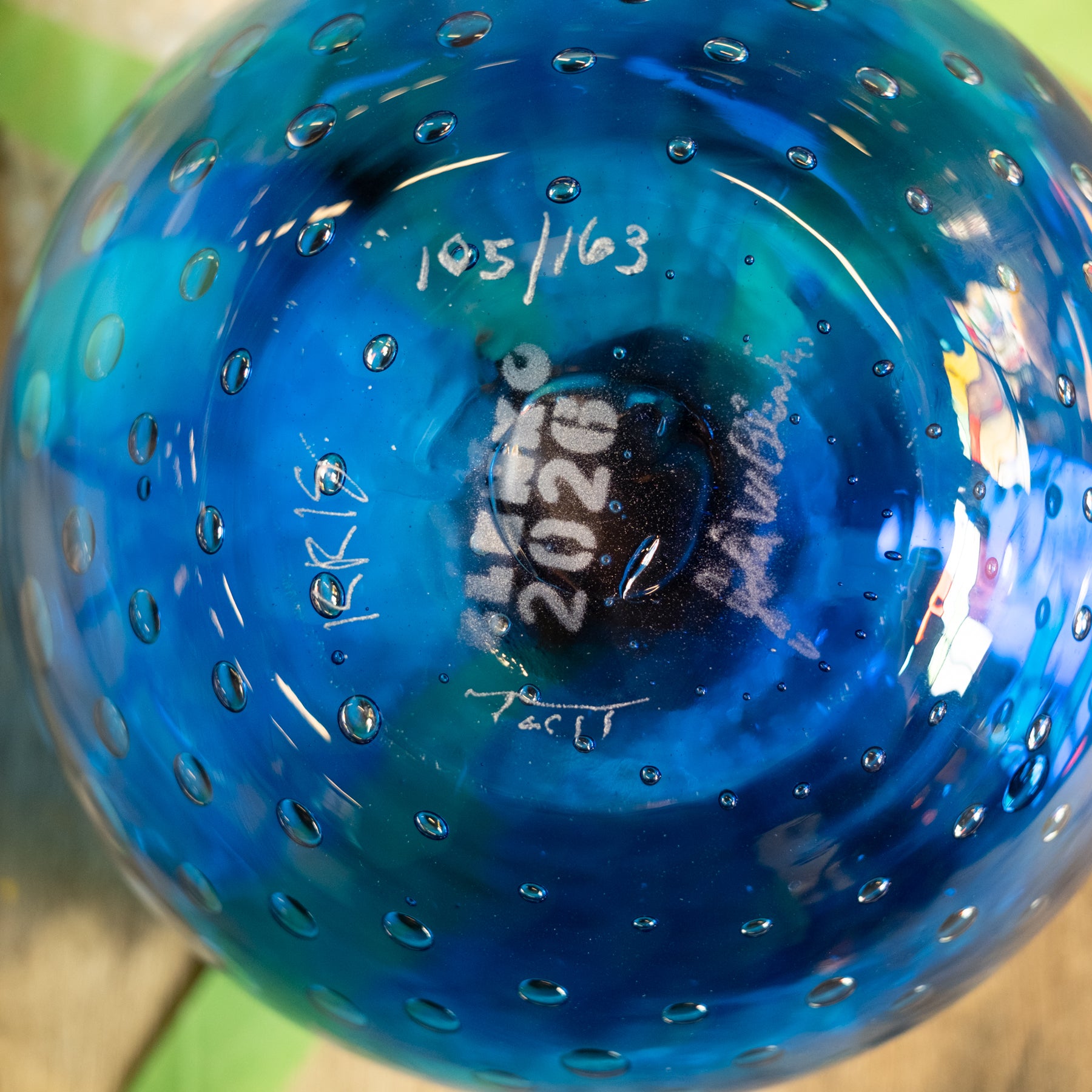 Close-up of the bottom of a translucent blue glass bowl with a bubble pattern, showing hand-written markings including '105/163' and '2020'.