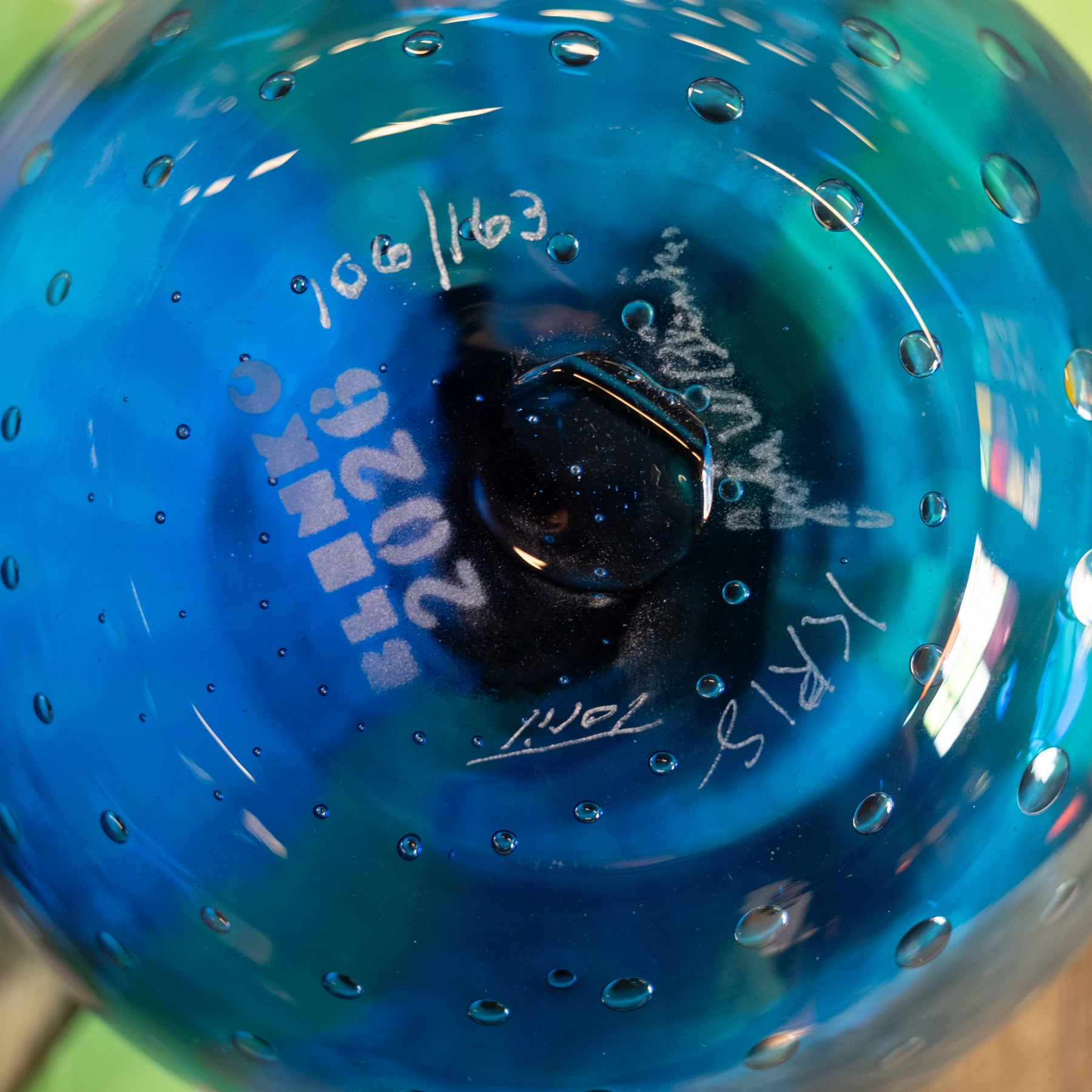 Close-up of the bottom of a blue glass vessel with a bubble pattern, showing white hand-written markings including '2024', '100/163', and 'SKX'.