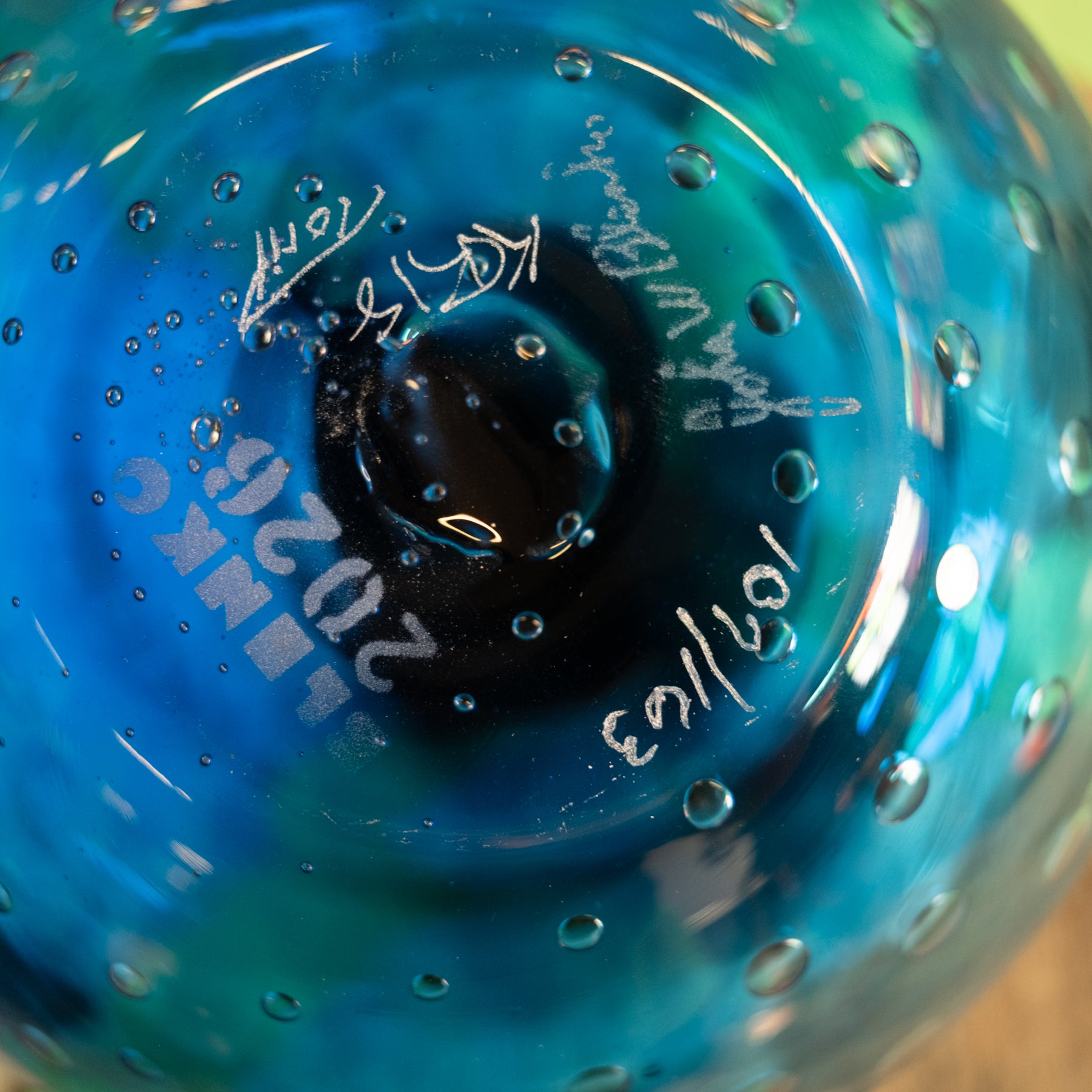 Close-up of a blue and turquoise hand-blown glass object with a bubbled texture and white handwritten markings inside the bubble.