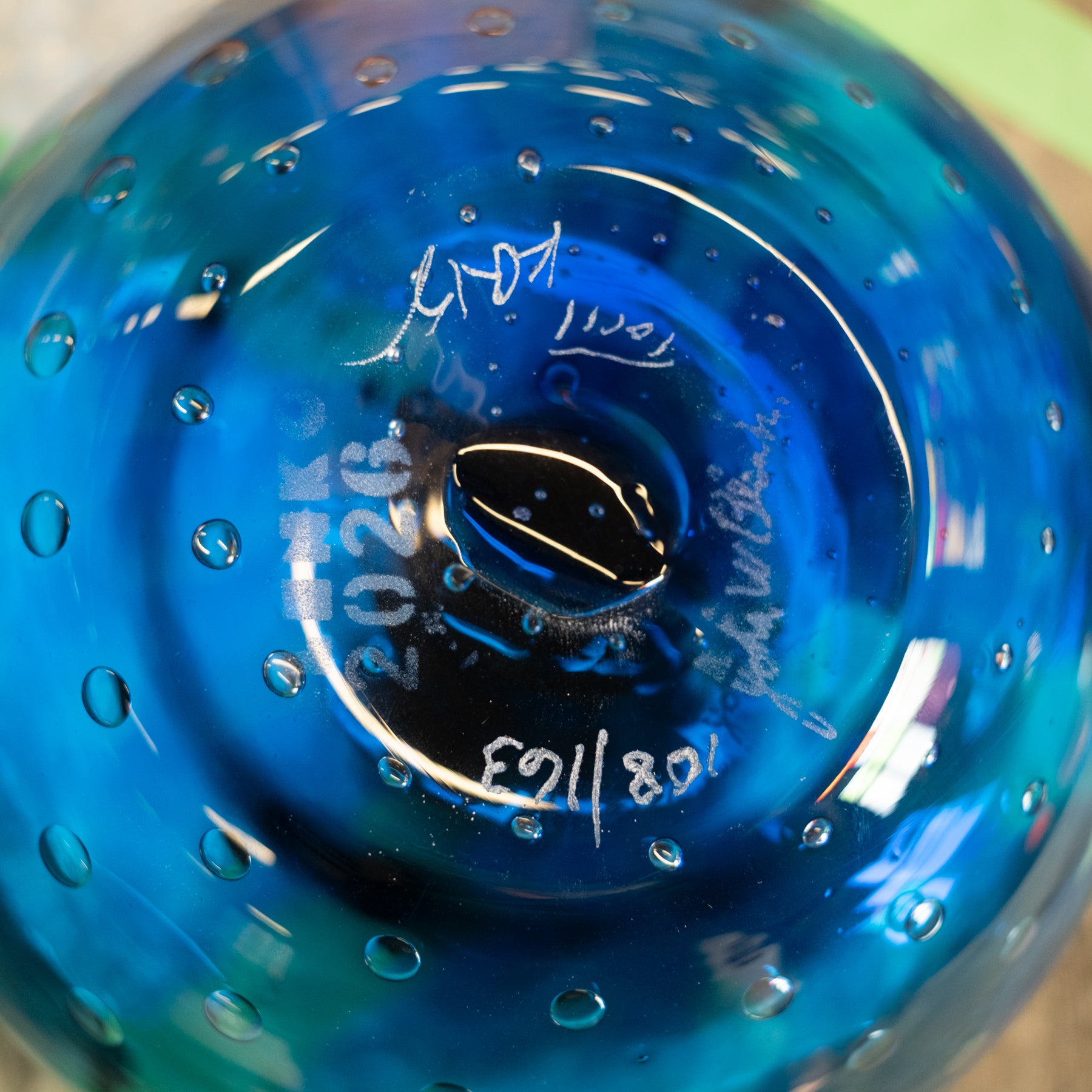 Close-up of the bottom of a blue glass bowl or vase with a bubble pattern, featuring white hand-written maker's marks and dates.
