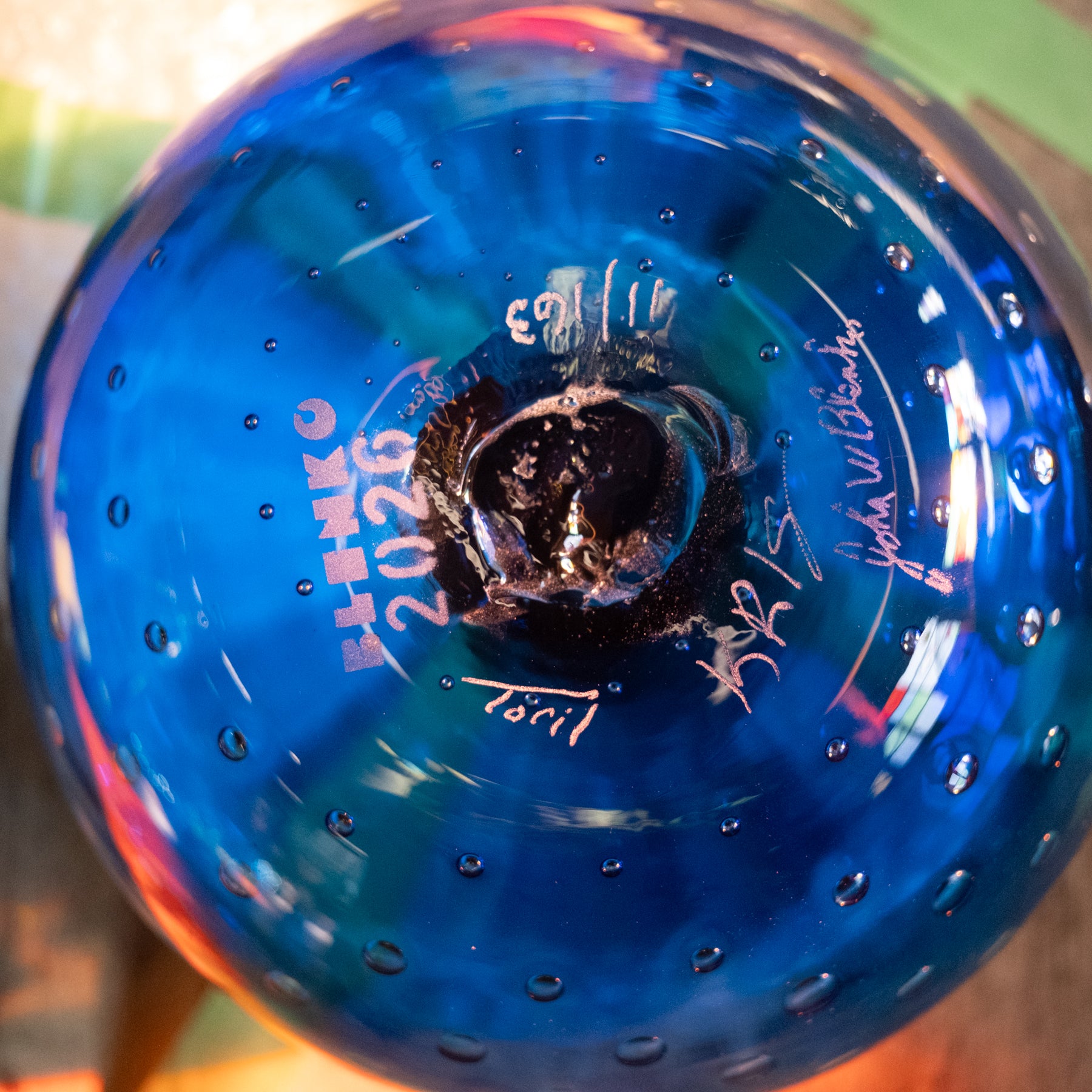 A close-up of a blue glass bottle or vase viewed from the bottom, showing a dark central base, small bubble-like perforations around the surface, and white printed text and signatures including 'SHIKO 2026' and 'Tori'.