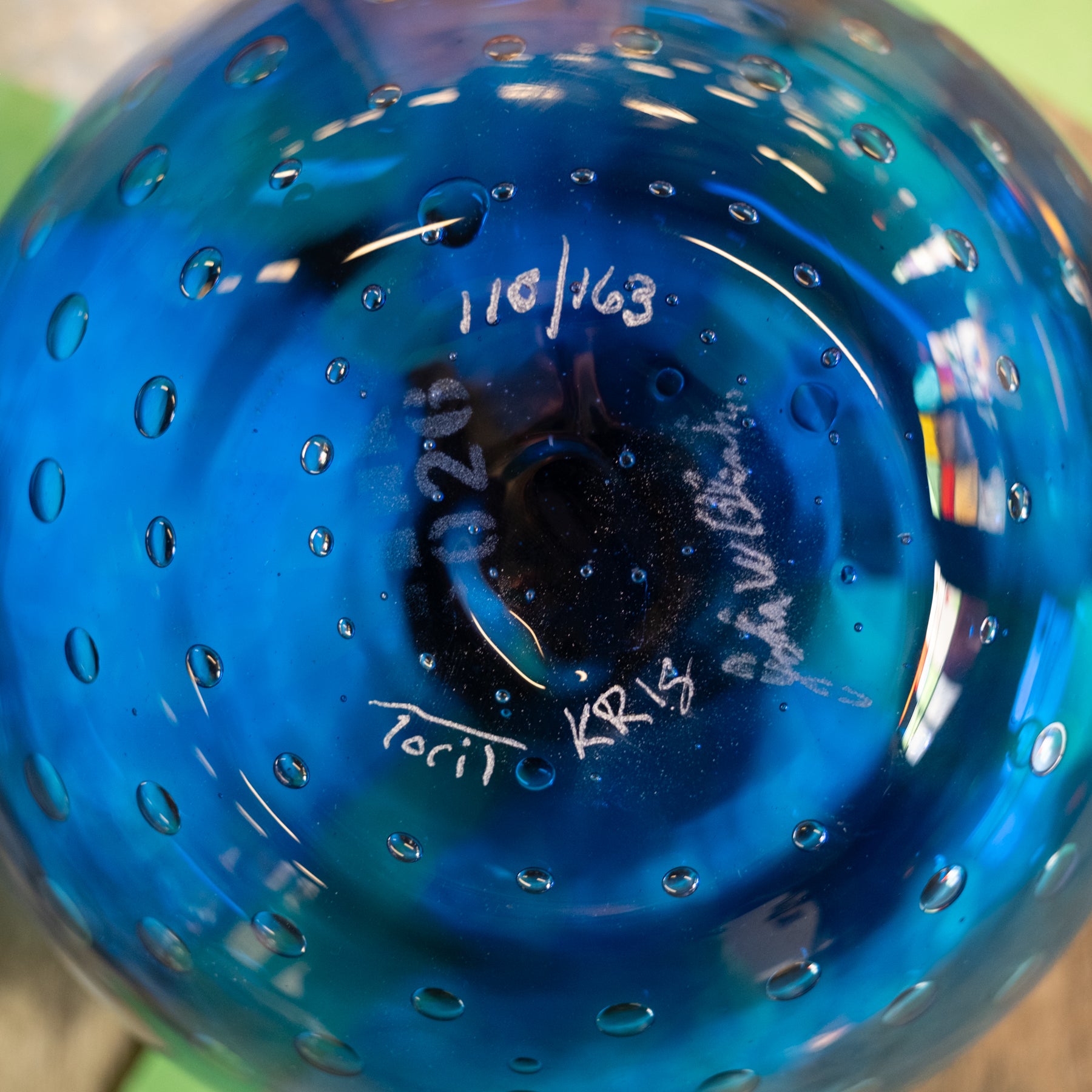 Close-up of the bottom of a blue glass vessel with a perforated bubble pattern, showing white handwritten markings and reflections.