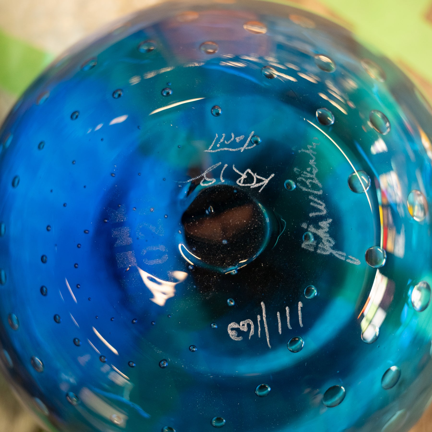 Close-up of the bottom of a hand-blown glass bowl or vase in vibrant blue with white hand-written maker's marks and serial numbers.