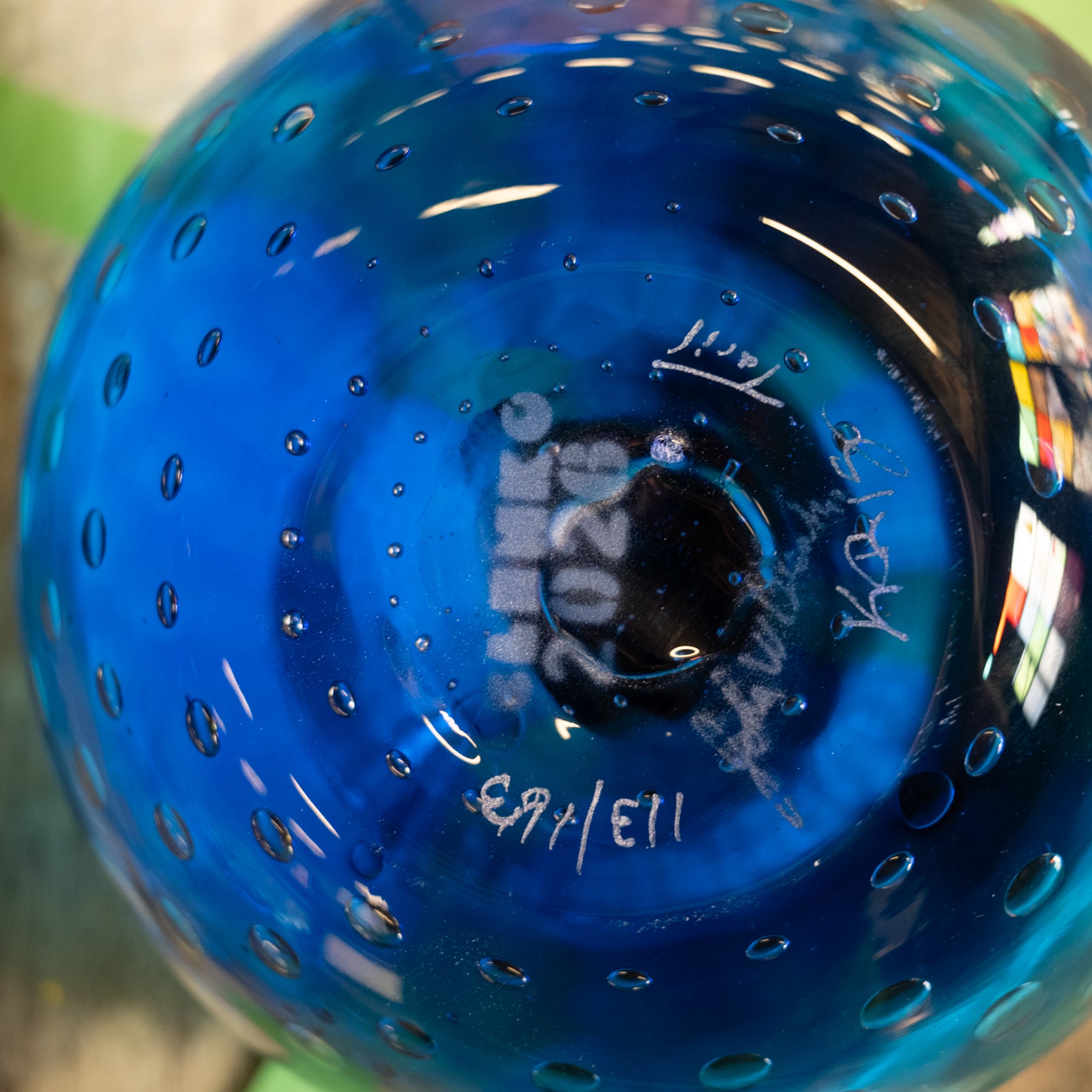 Close-up of a blue glass bowl or vase with a bubbled texture, featuring white handwritten markings and signatures on the bottom.
