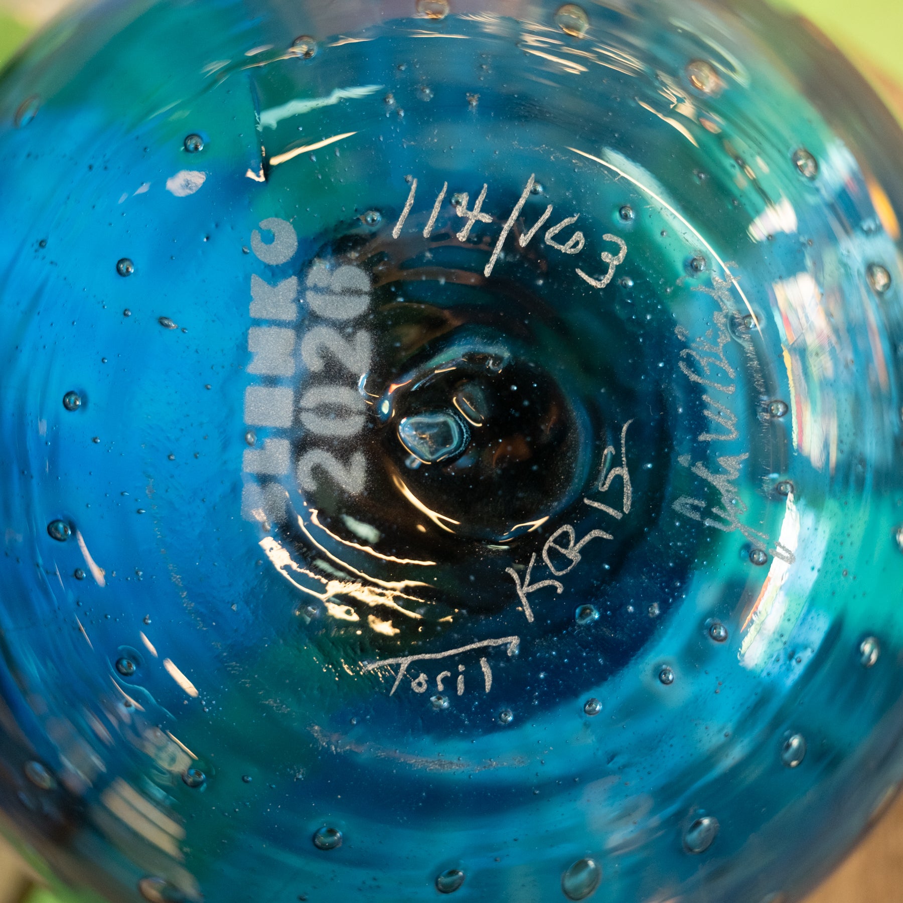 Close-up of the bottom of a blue glass bowl or vase with water droplets, showing etched maker's marks including 'SHENKO 2026' and handwritten dates and signatures.