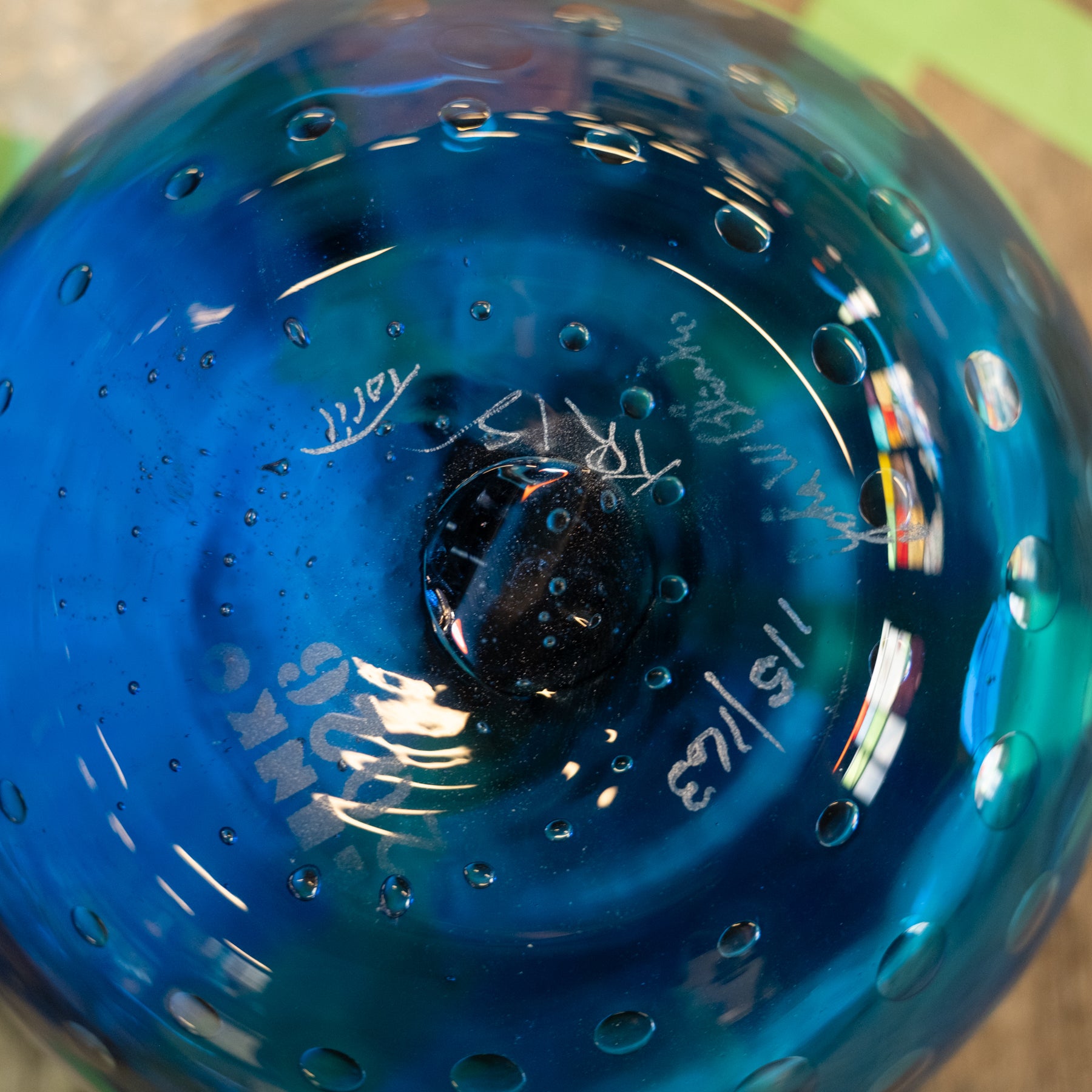 Close-up of a blue glass bowl with etched markings and bubble details, showing reflections and part of the maker's signature.