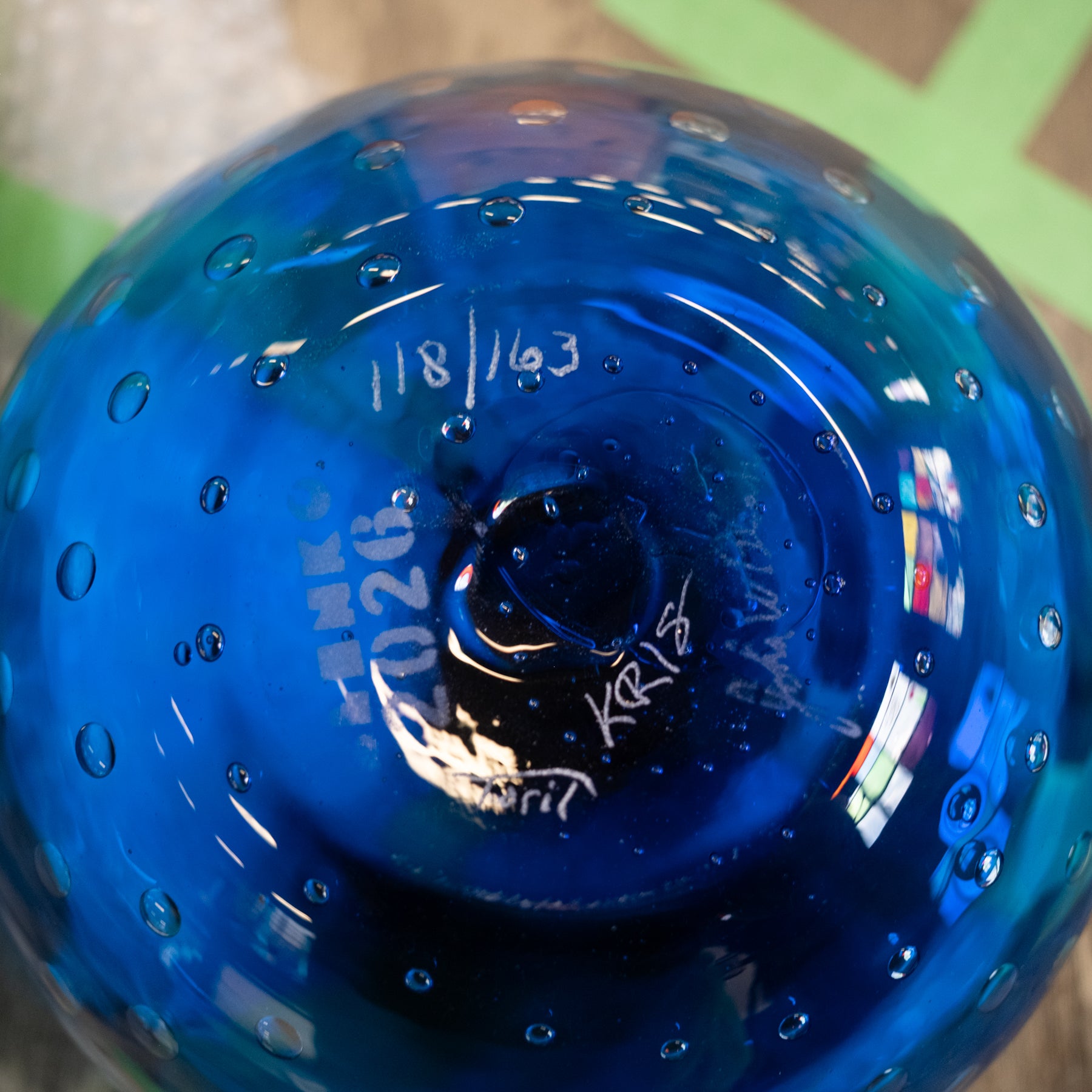 A close-up of a blue glass bowl or vase showing the bottom of the vessel, with white handwritten markings and bubbles in the glass, photographed against a blurred background.