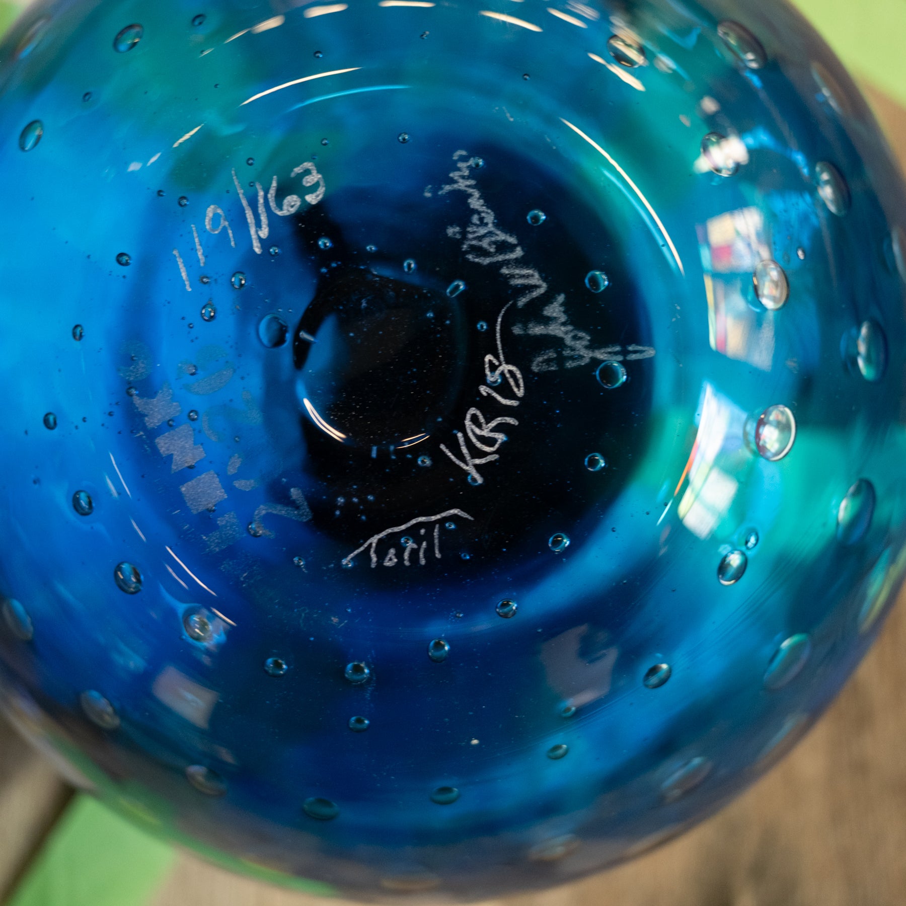 A close-up of a blue glass bowl or vase with a textured bubble pattern and white hand-written markings on the bottom.
