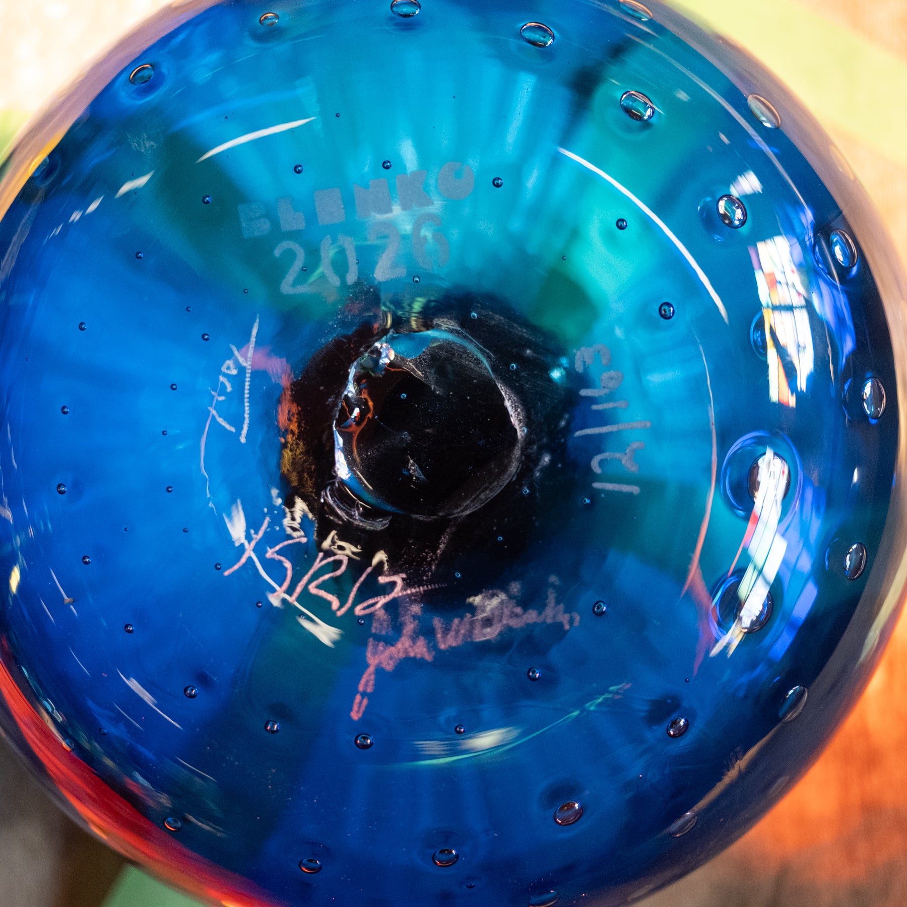 A close-up of a round blue glass object, likely a decorative bowl or vase, showing a textured blue surface with small bubbles, a dark central hole, and white handwritten markings including the date '2020' and signatures.