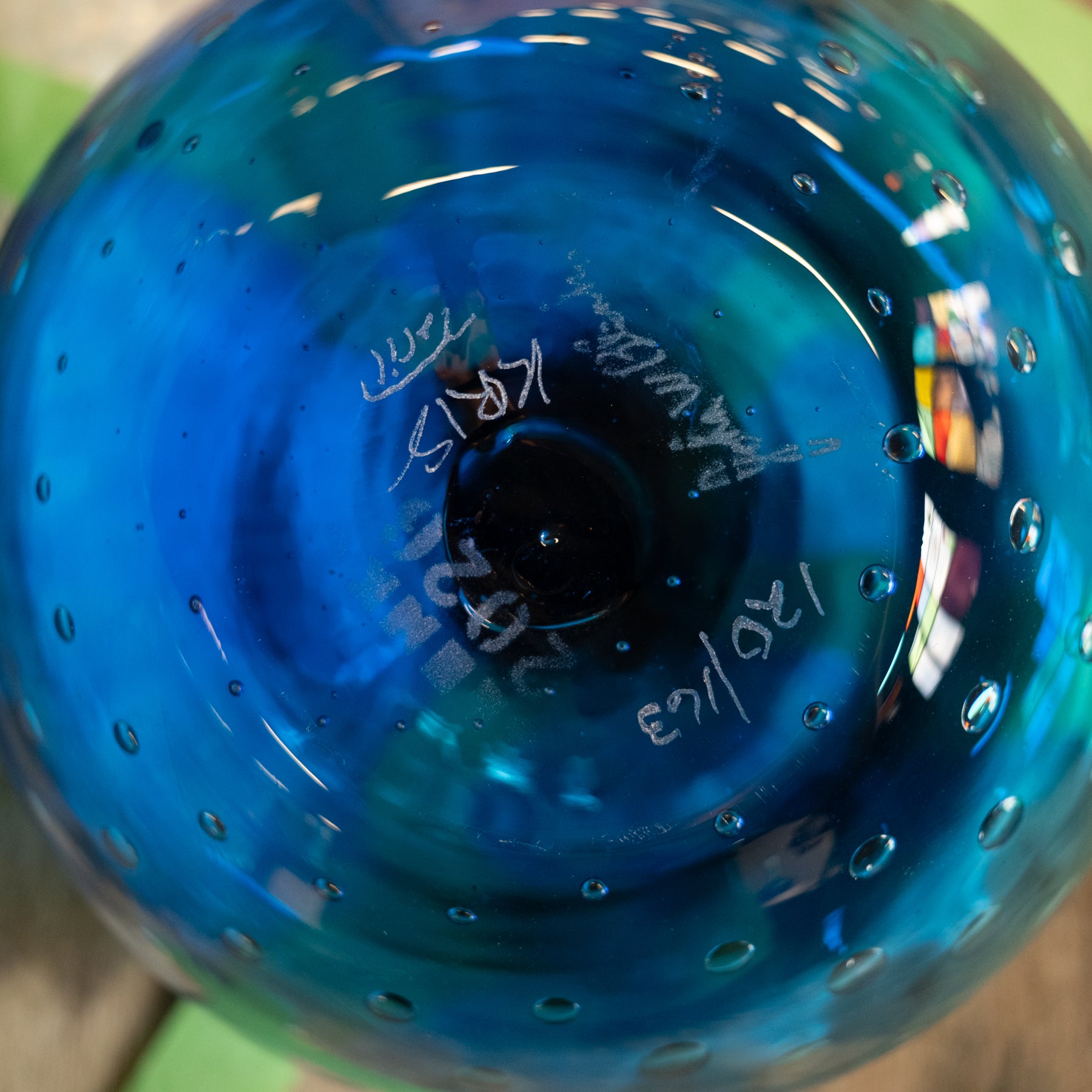 A close-up of a blue glass vessel, likely a vase or decorative bowl, showing a textured spiral pattern with small bubble details and white handwritten markings near the center.