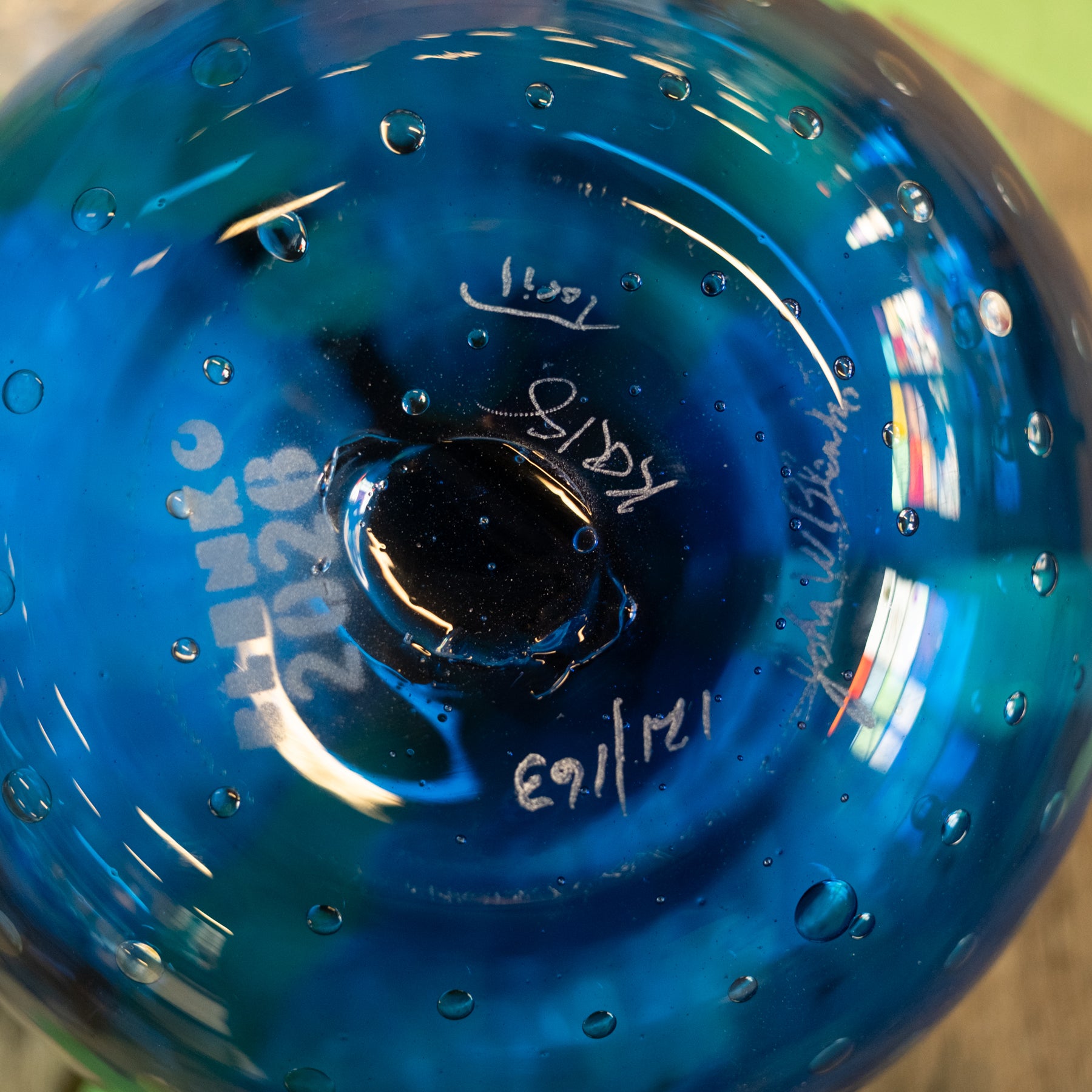 Close-up of the bottom of a transparent blue glass container or bottle, showing water droplets on the surface and white handwritten markings.