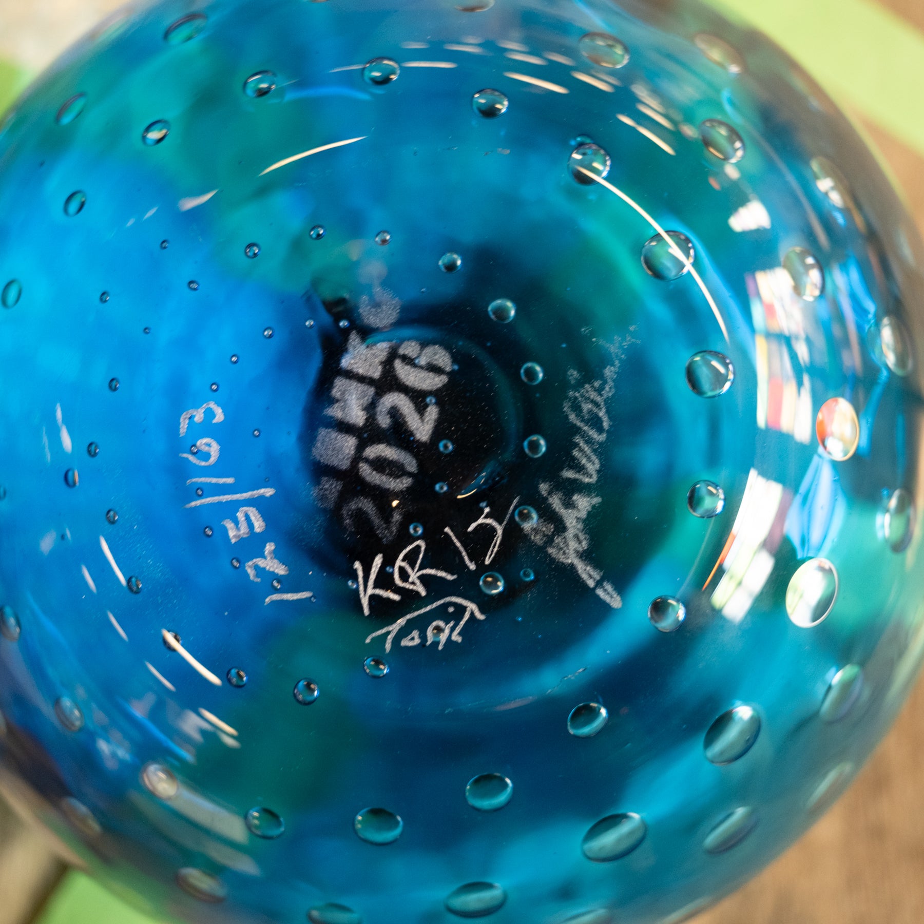 Close-up of a blue glass bowl or vase with a bubbled texture and white handwritten markings inside, photographed against a softly blurred background.